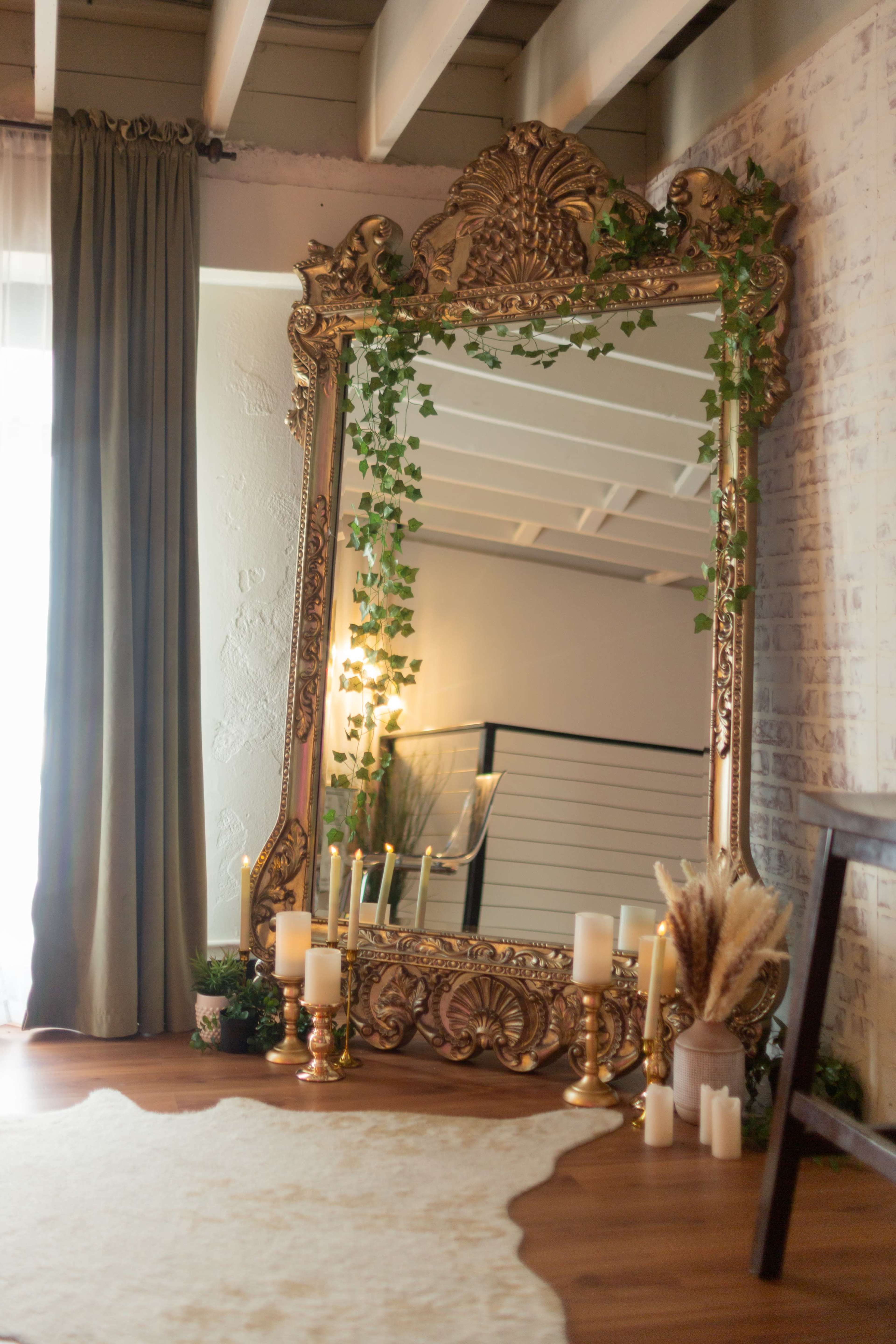 A large ornate mirror with a decorative frame is placed against a wall, surrounded by green plants and candles on the floor.