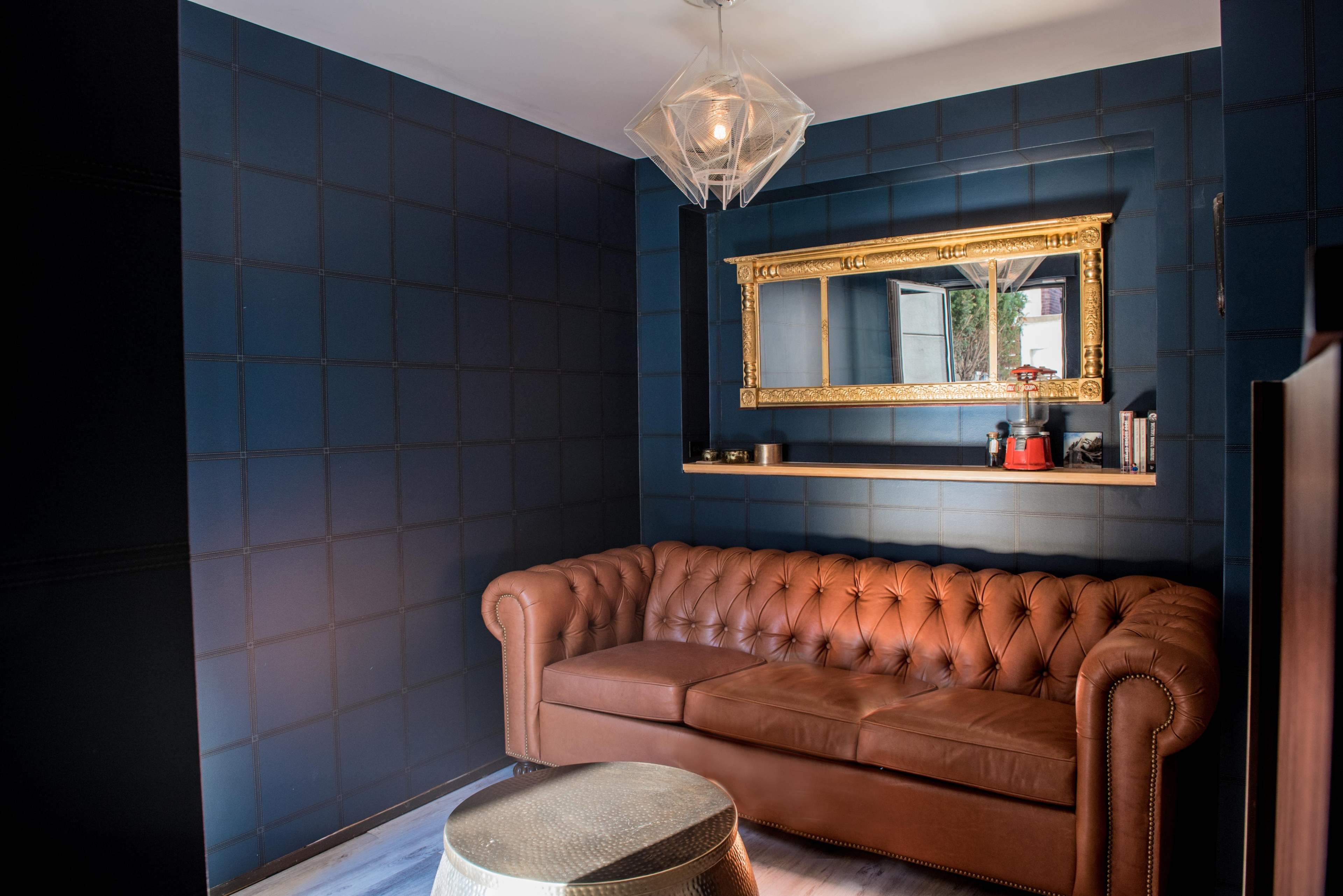 A modern room with a dark blue wall, a tufted brown leather couch, a decorative mirror above a shelf, and a round metallic coffee table.