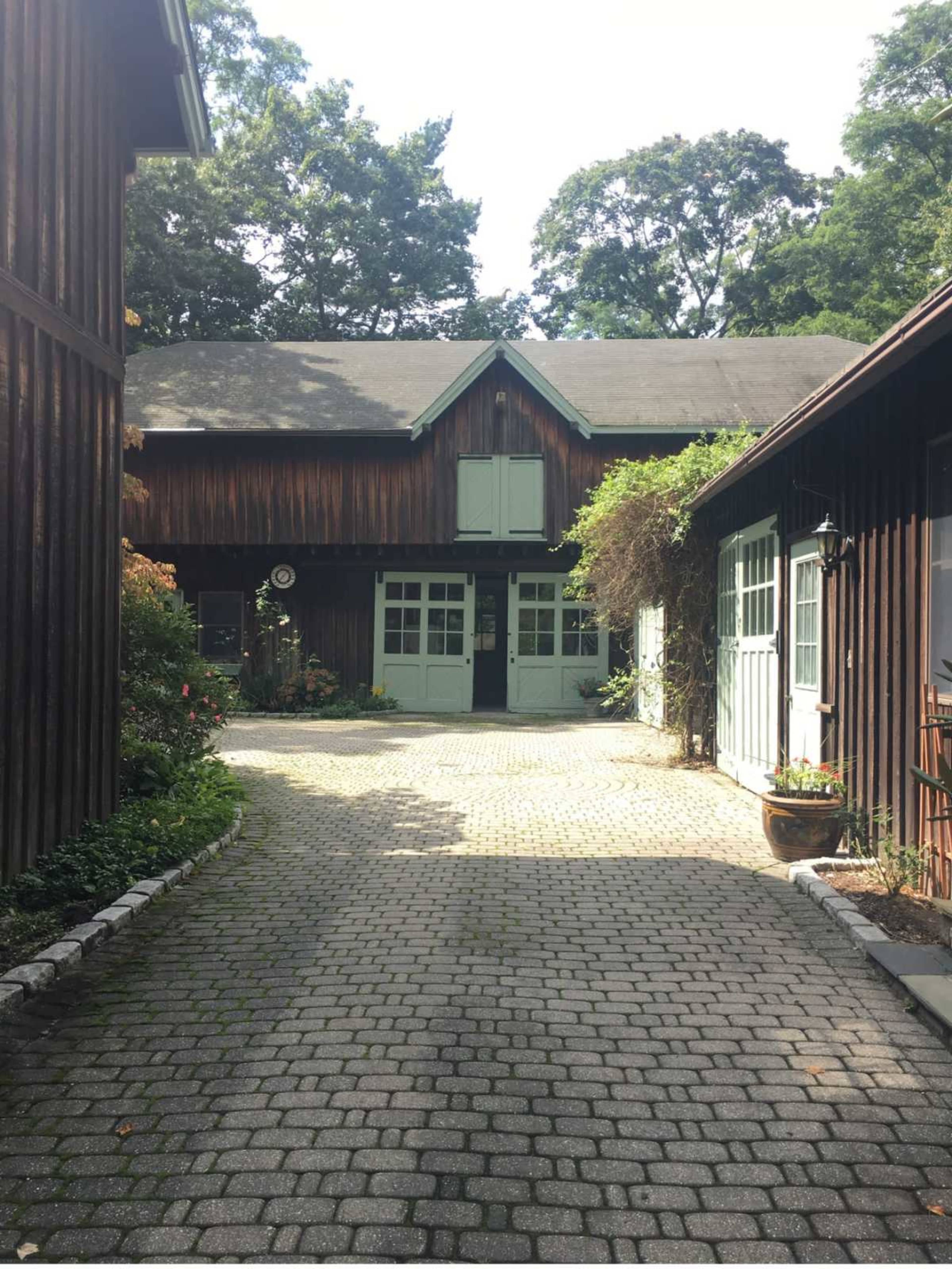 The image shows a cobblestone pathway leading to a large wooden building, surrounded by greenery and trees.