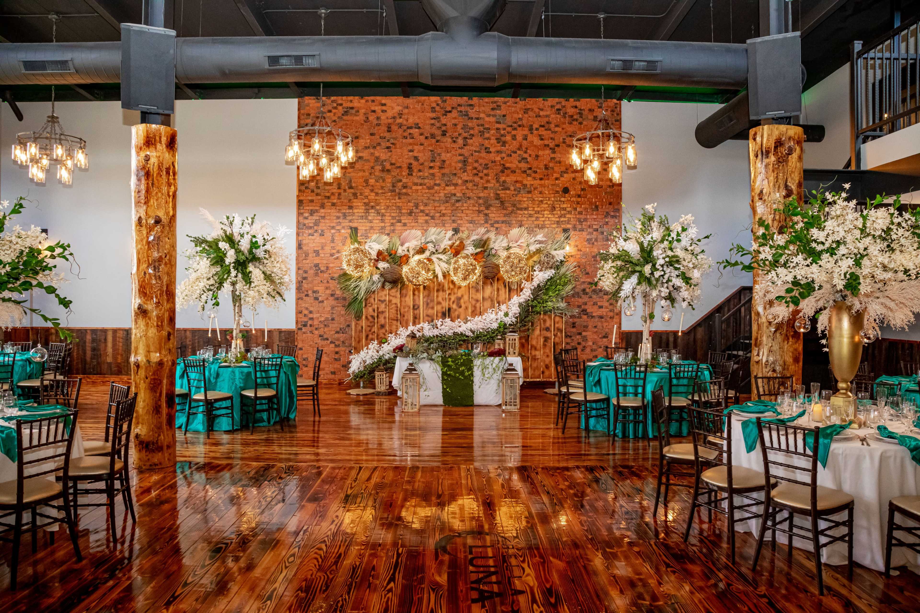 An elegantly decorated event space featuring tables with turquoise cloths, large floral arrangements, and a backdrop of greenery and flowers against a brick wall.