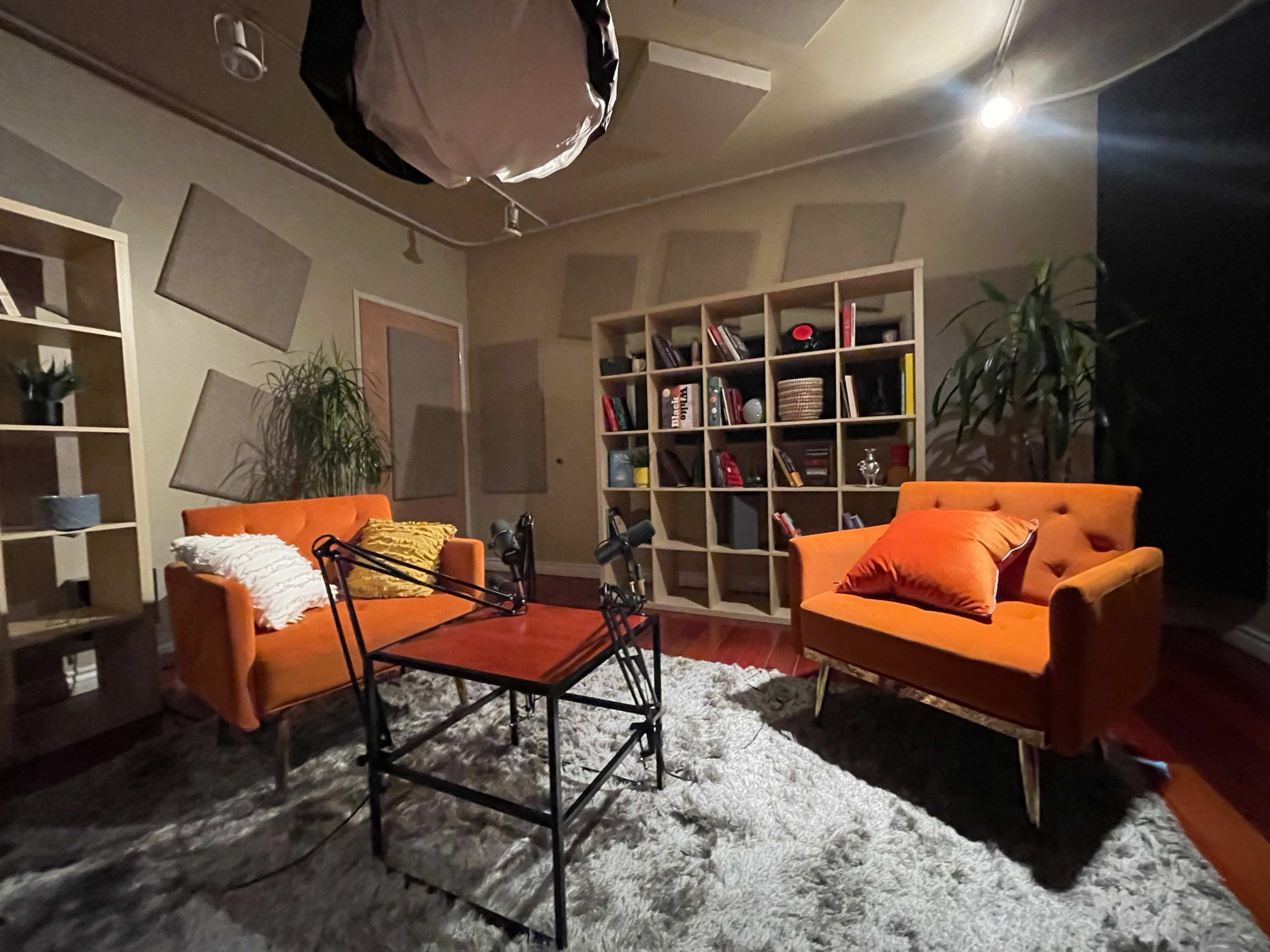 The image shows a cozy studio setup with two orange sofas, a wooden coffee table, and a bookshelf filled with various items, all arranged on a plush gray rug.