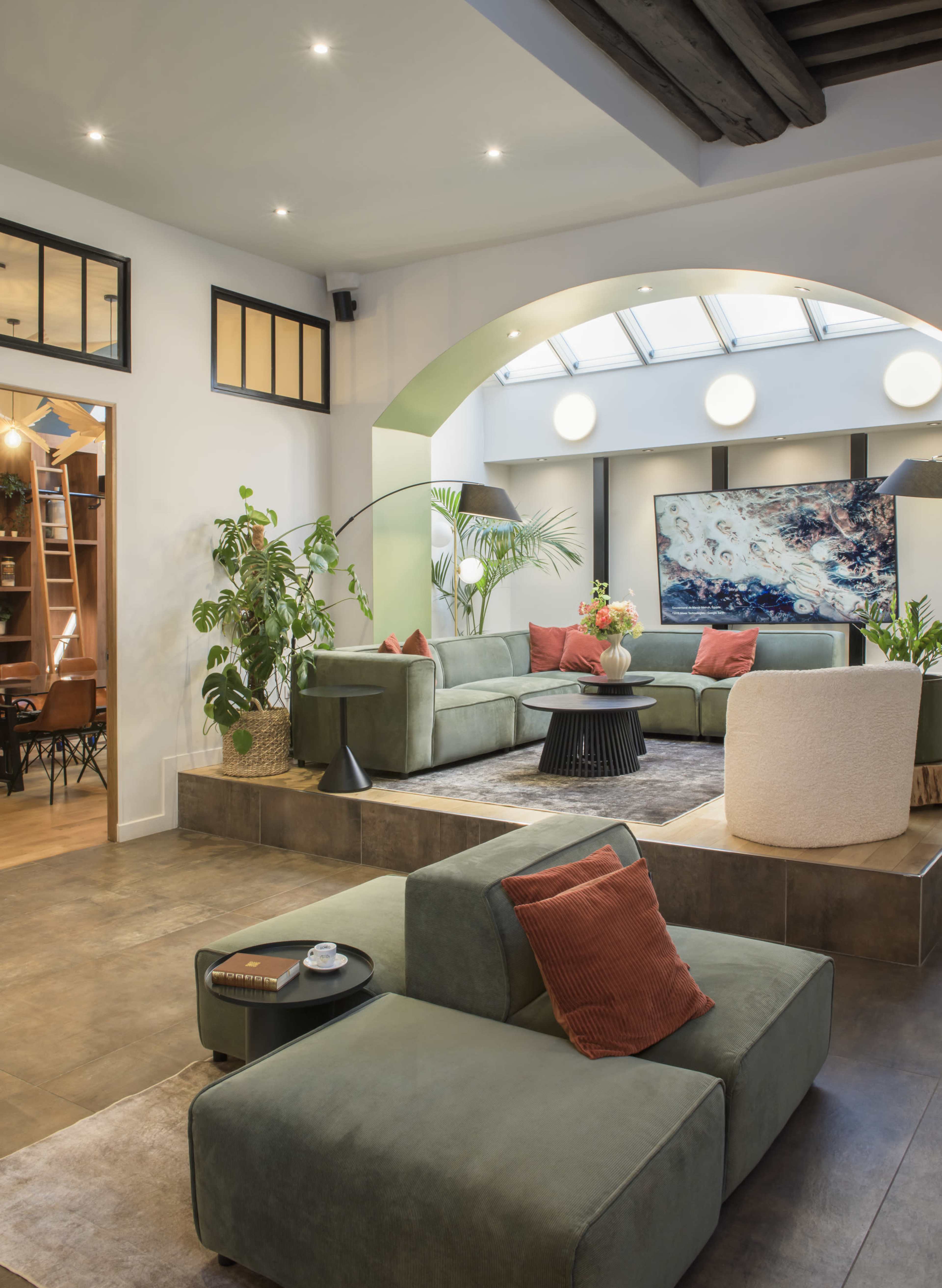 The image shows a modern, spacious living area featuring green sofas, indoor plants, and a large artwork illuminated by natural light from a skylight.