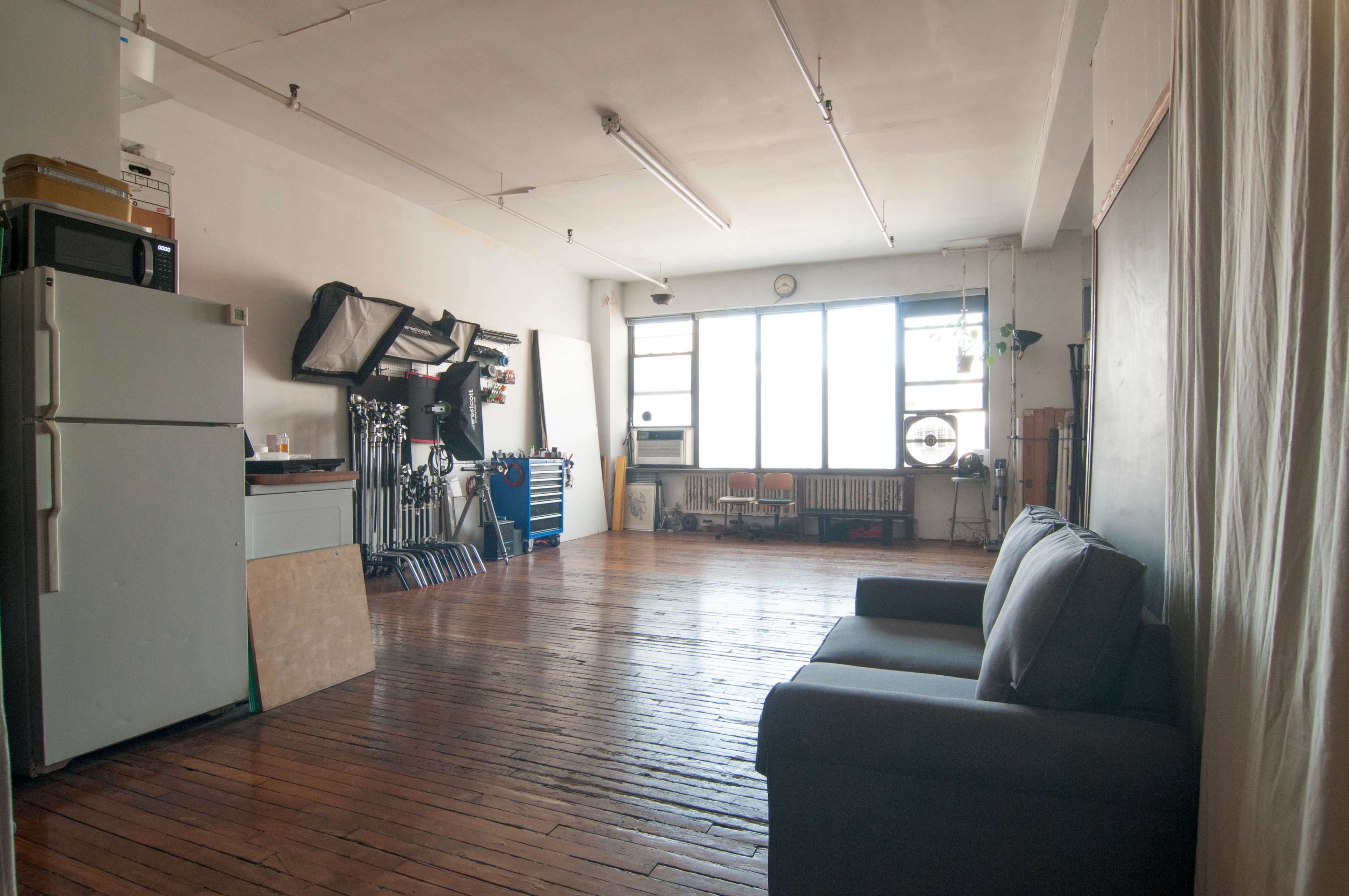 The image shows a spacious, vacant room with wooden floors, a sofa, a large window, and various equipment against the walls.