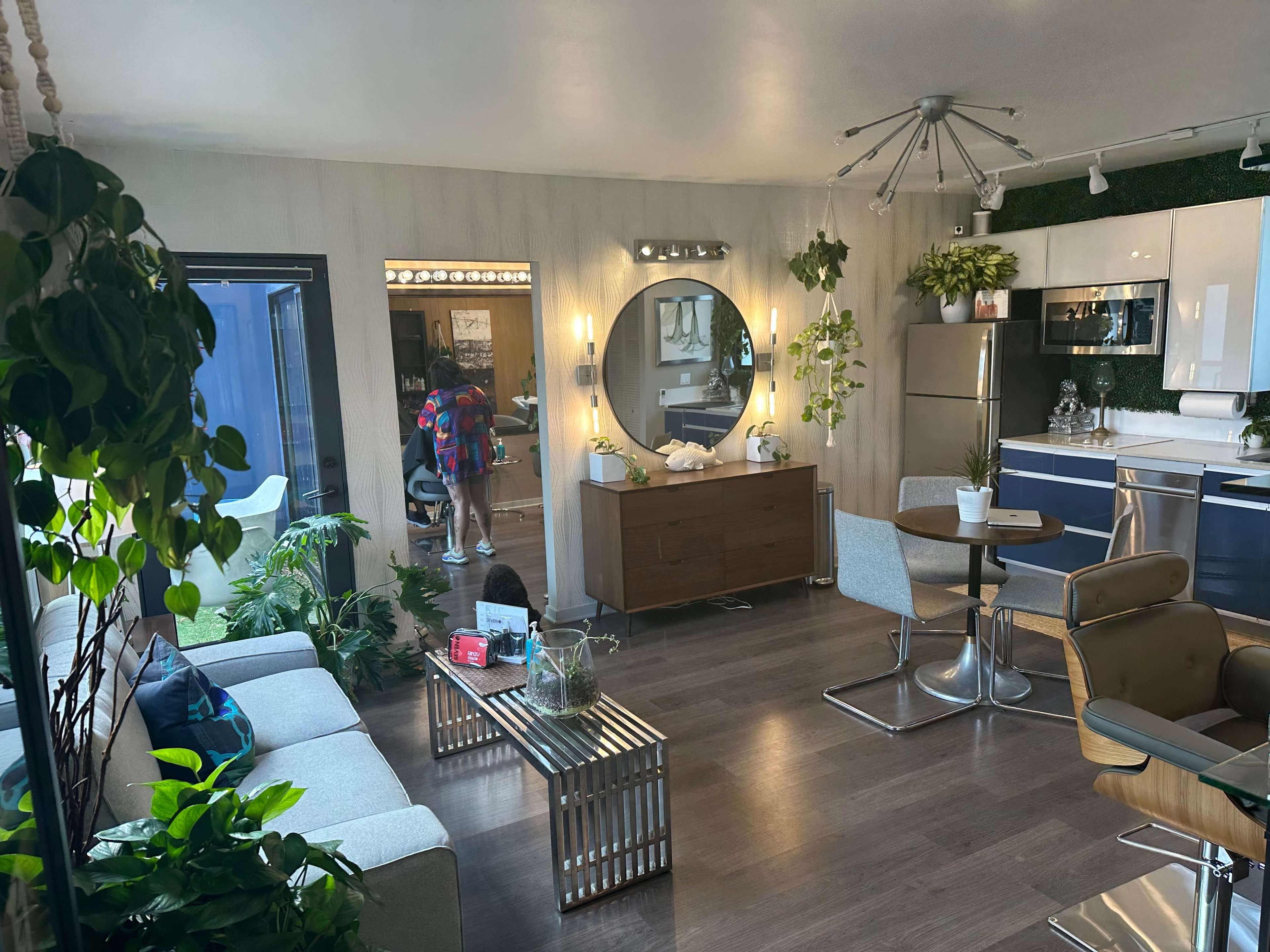 A modern living space with a sofa, wooden cabinet, and a kitchen area, surrounded by green plants and natural light.