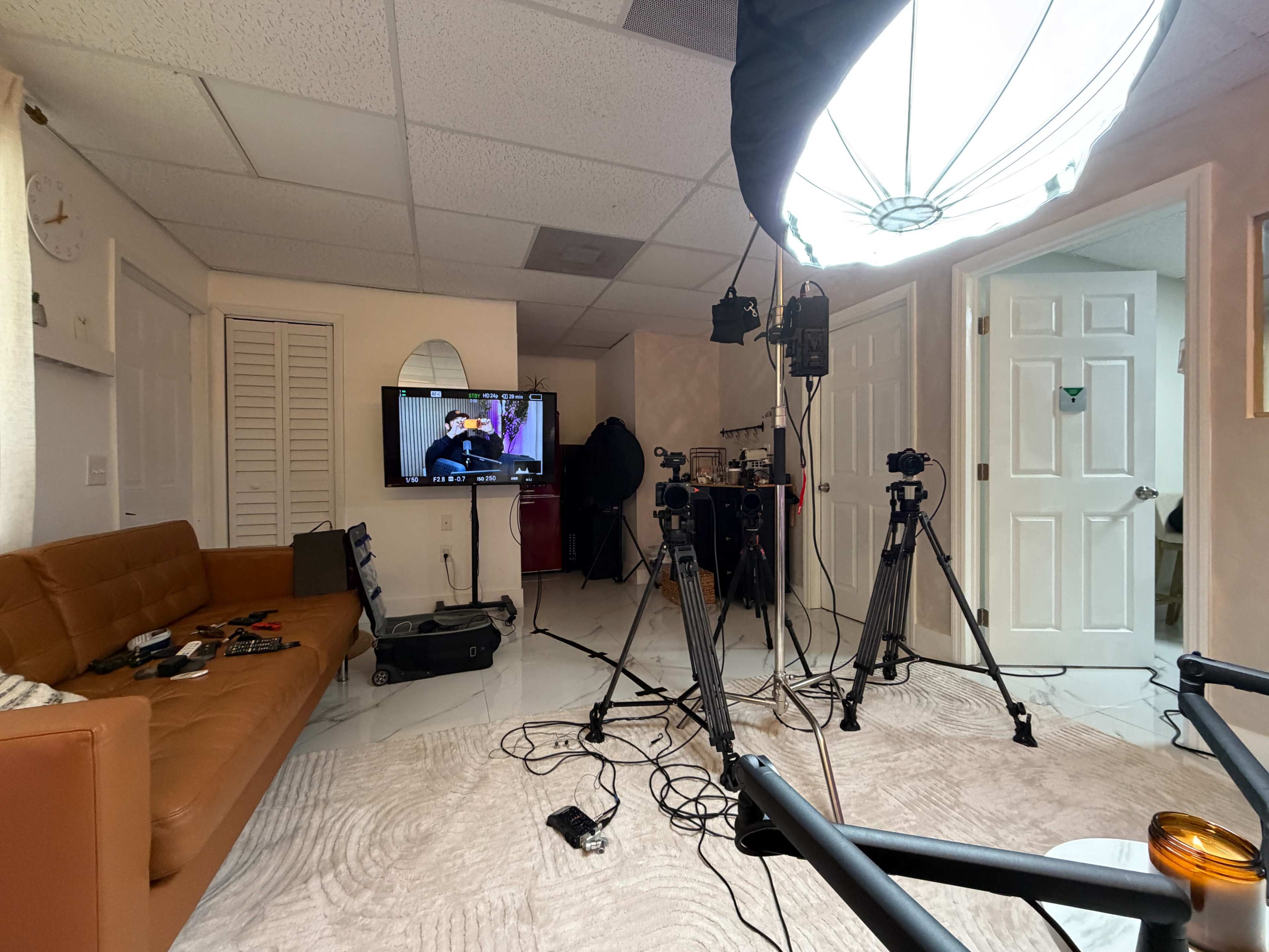 The image shows a room setup for filming, featuring a brown couch, a TV displaying a program, two tripods with cameras, and various equipment on the floor.