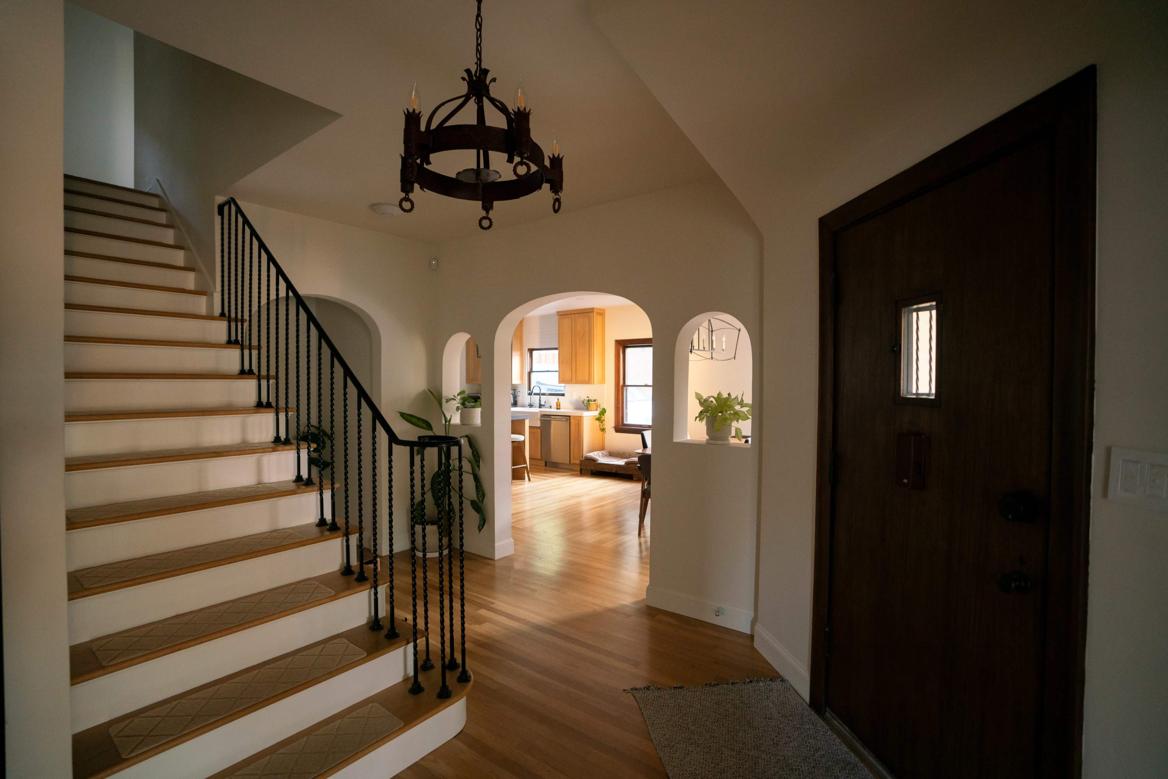 The image shows a well-lit entryway featuring a staircase with a decorative railing, an archway leading to a kitchen area, and a wooden front door.