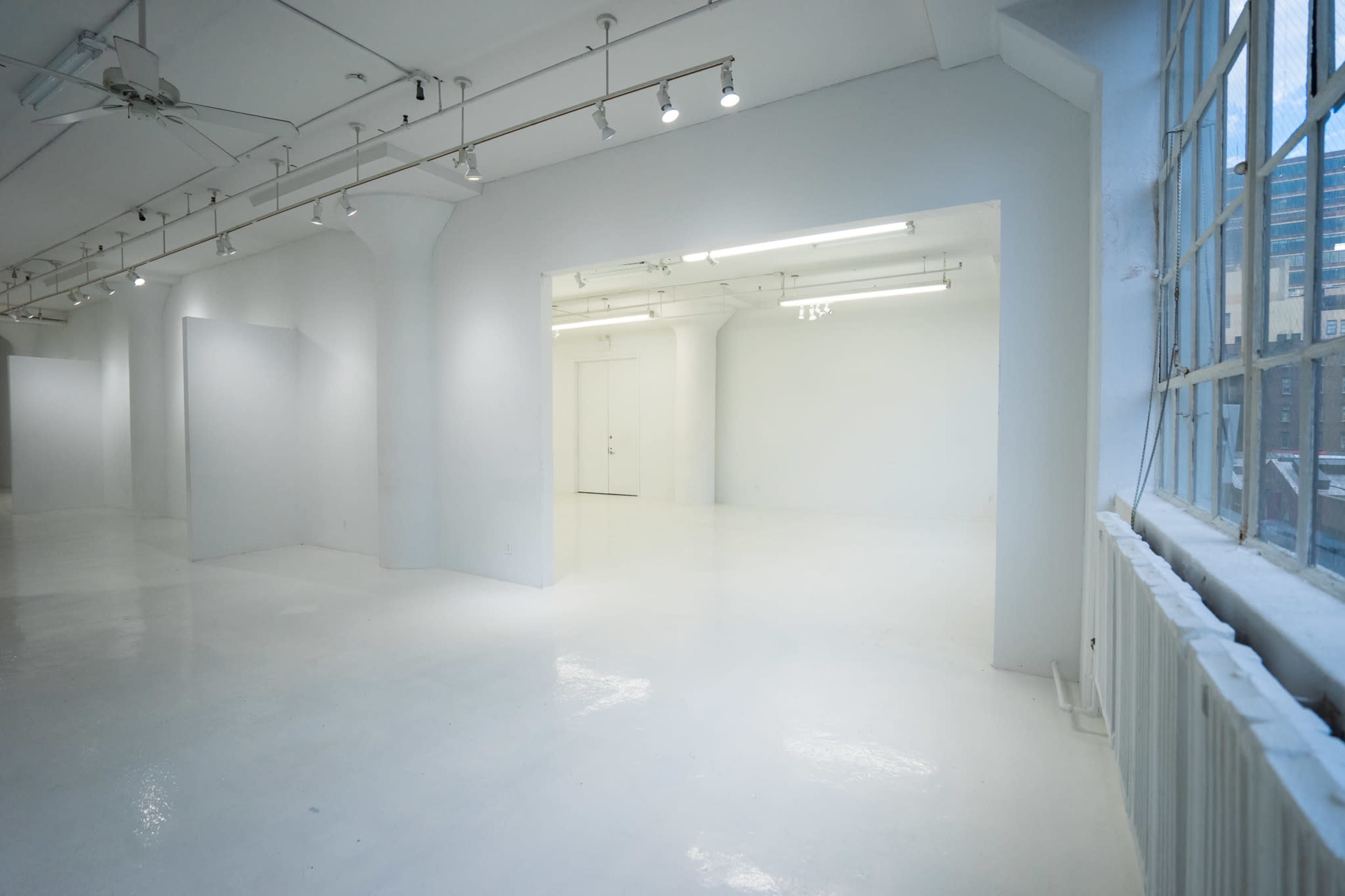 A spacious, empty gallery with white walls and a polished floor, featuring large windows that provide natural light.