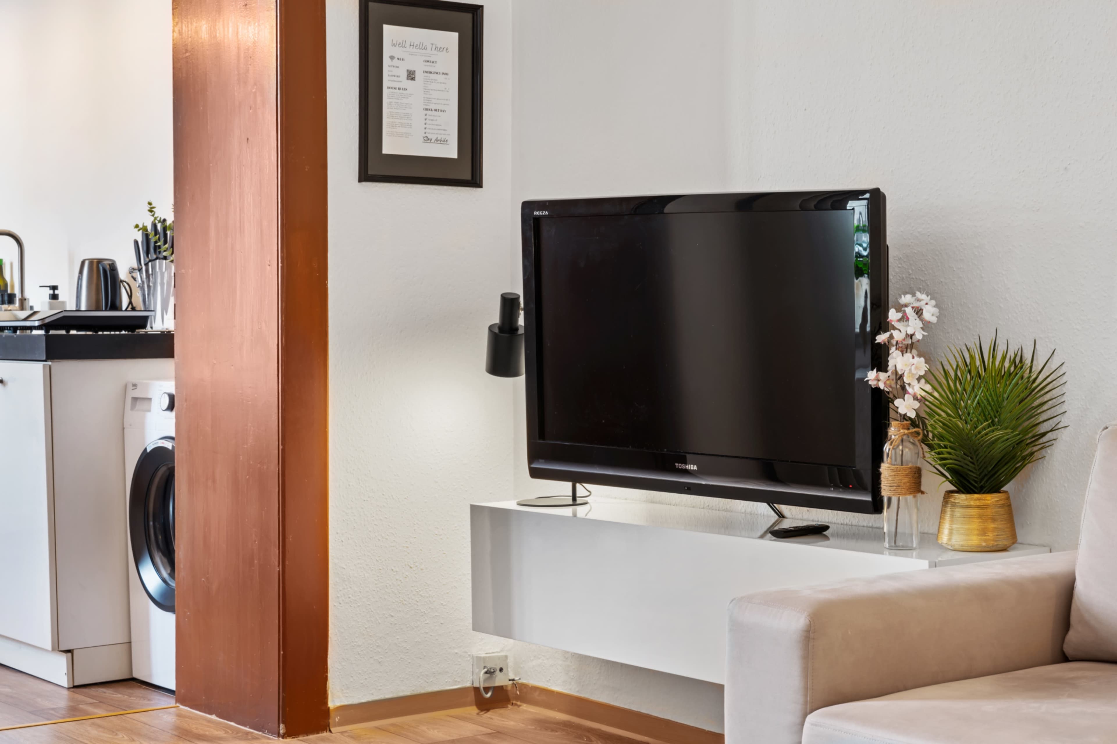The image shows a living space with a flat-screen television mounted on a white shelf, alongside a sofa and a view of a kitchen area with appliances.