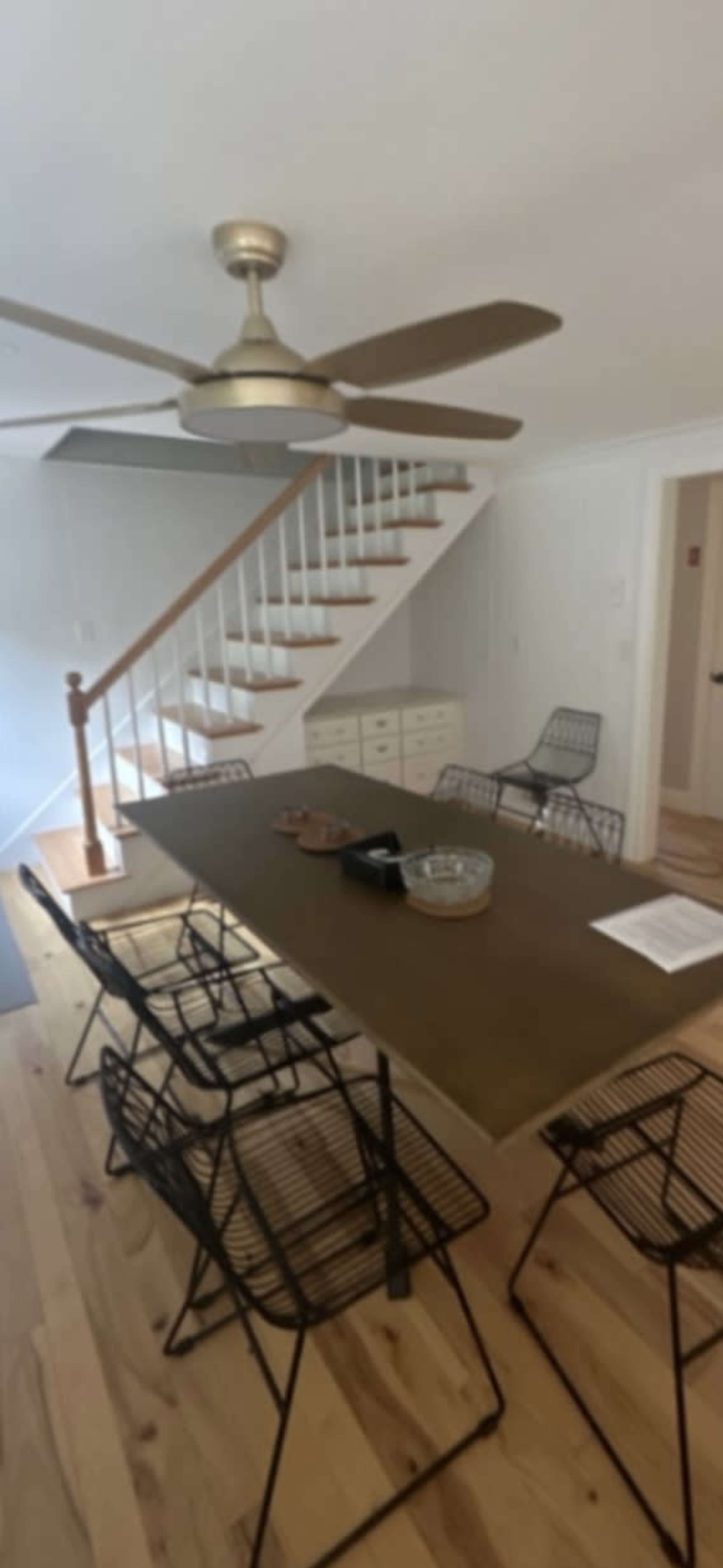 A dining area with a long table and six black wire chairs, adjacent to a staircase and well-lit by a ceiling fan.