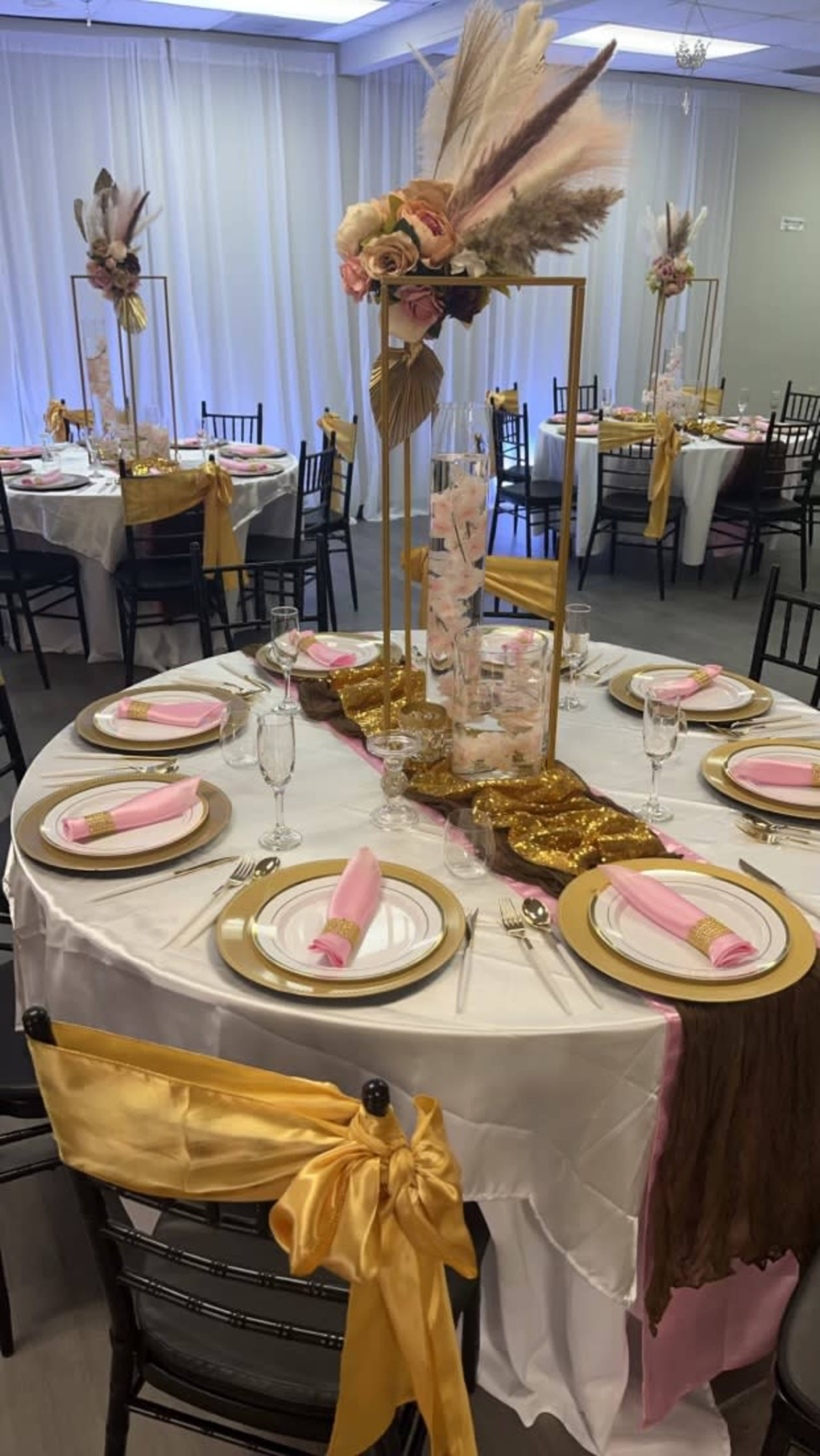 Elegantly arranged tables with gold and pink decorations, featuring tall floral centerpieces and folded napkins, set for a formal event.