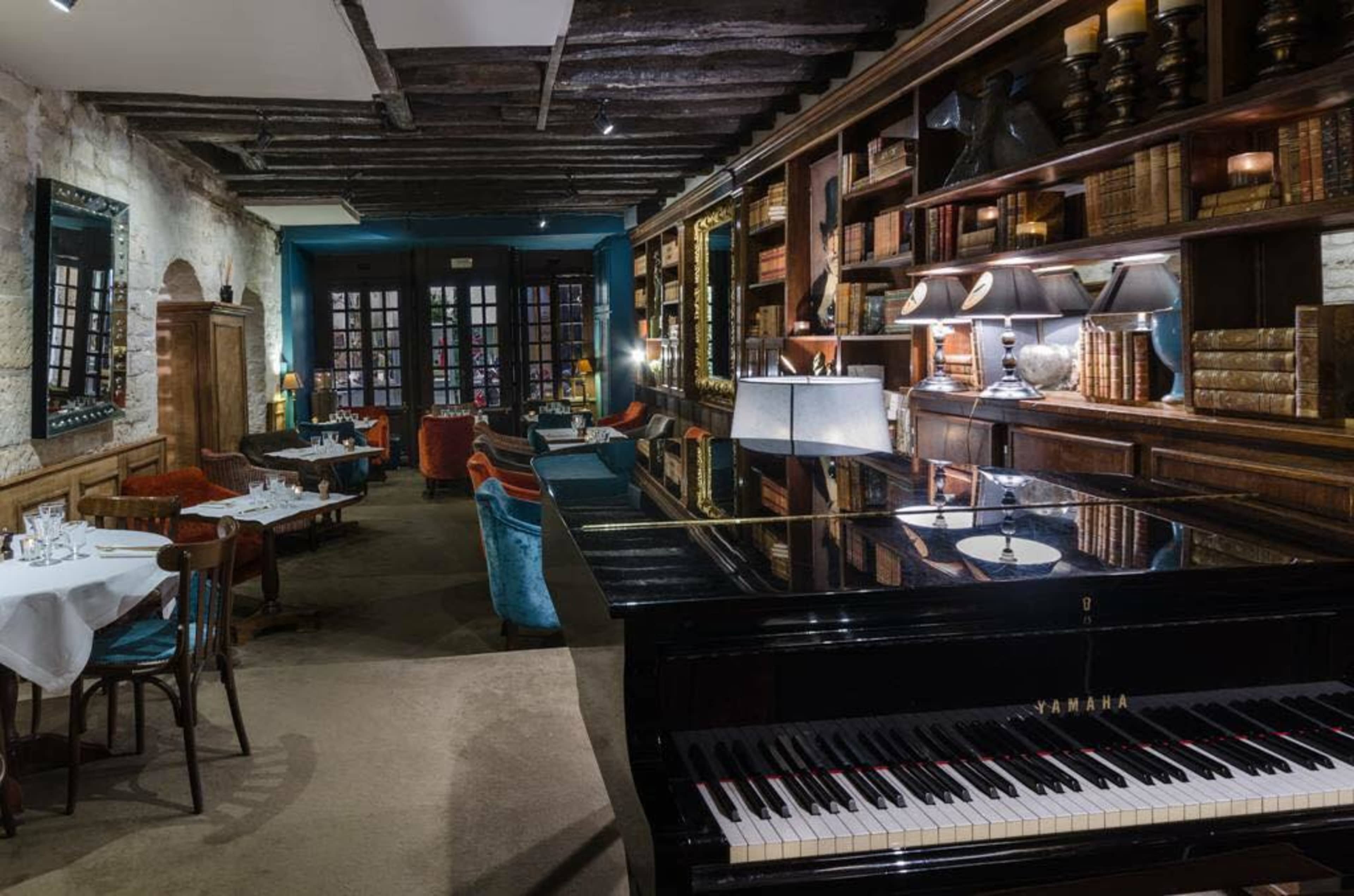 A black grand piano sits in a dimly lit restaurant filled with bookshelves, wooden beams, and table settings.