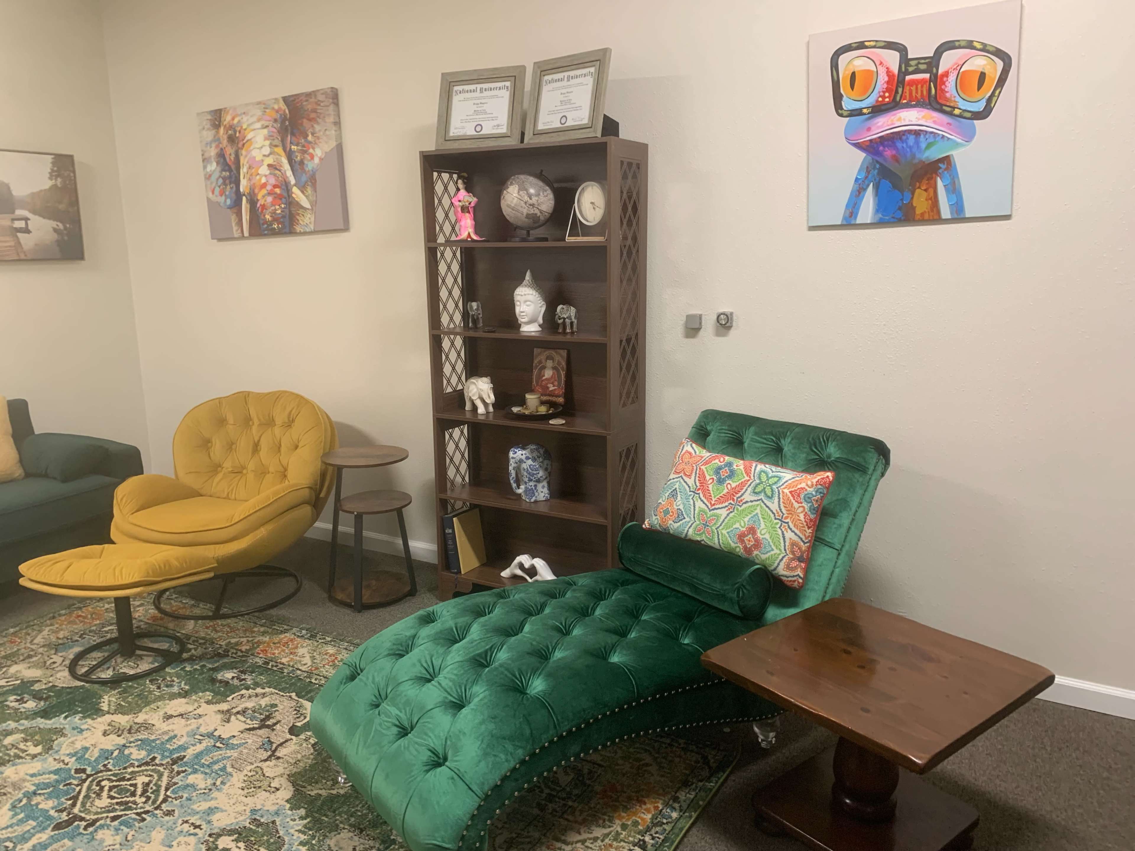 The image shows a cozy room with a green chaise lounge, a yellow accent chair, a wooden side table, and a bookshelf containing decorative items and framed certificates.
