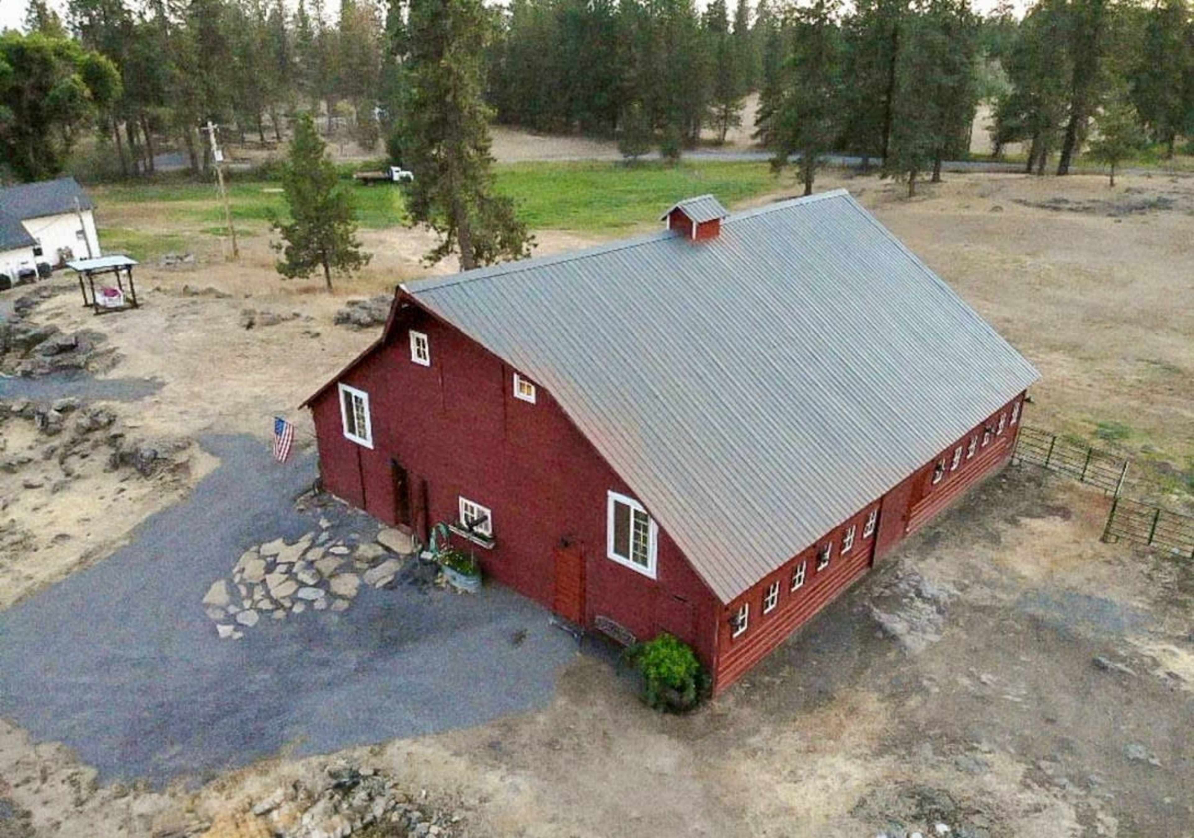 The image shows a large red barn with a metal roof situated on a dry landscape surrounded by trees.