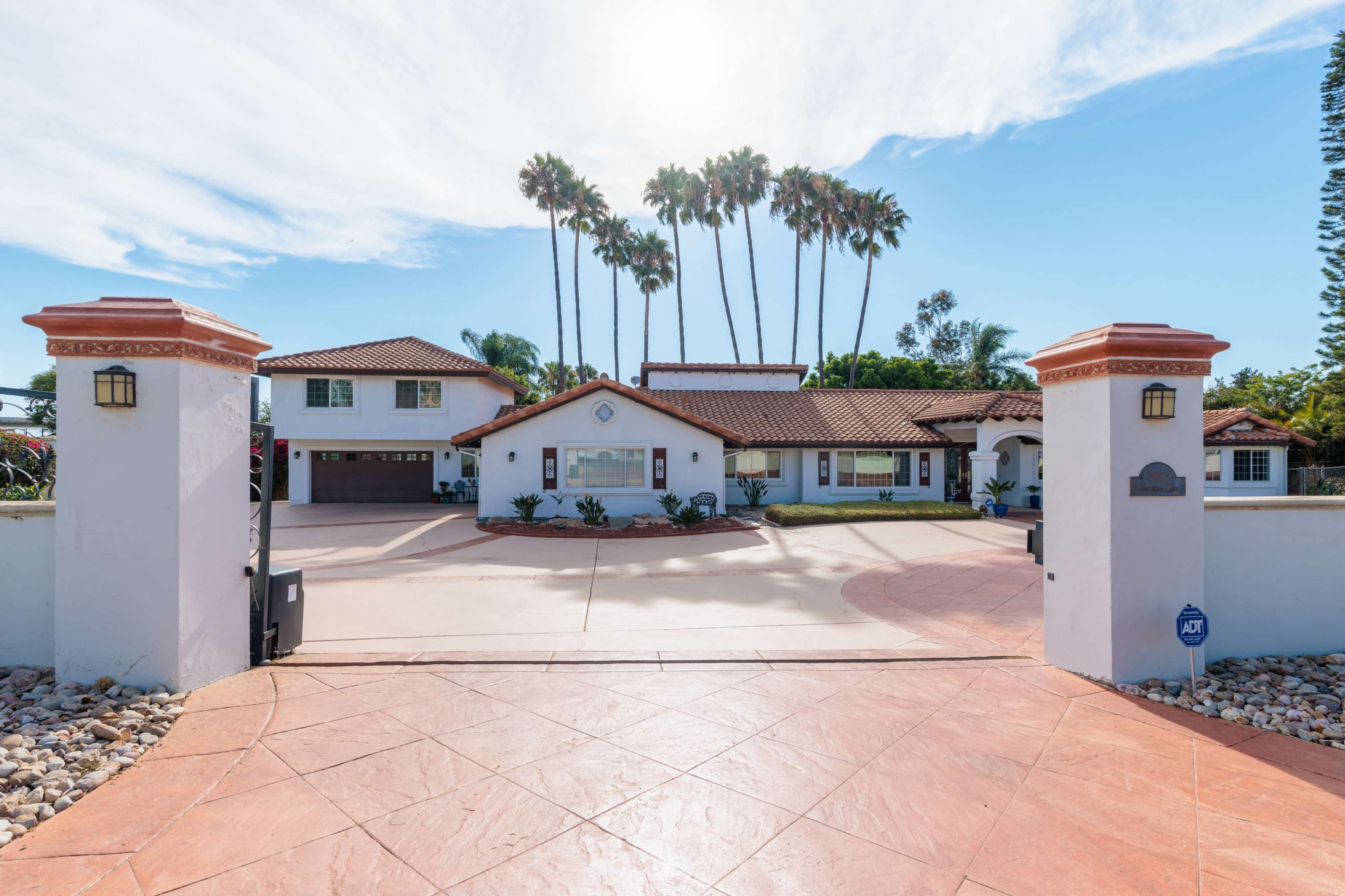 A large, two-story house with a tile roof is situated behind a gated entrance, framed by palm trees and surrounded by a paved driveway.