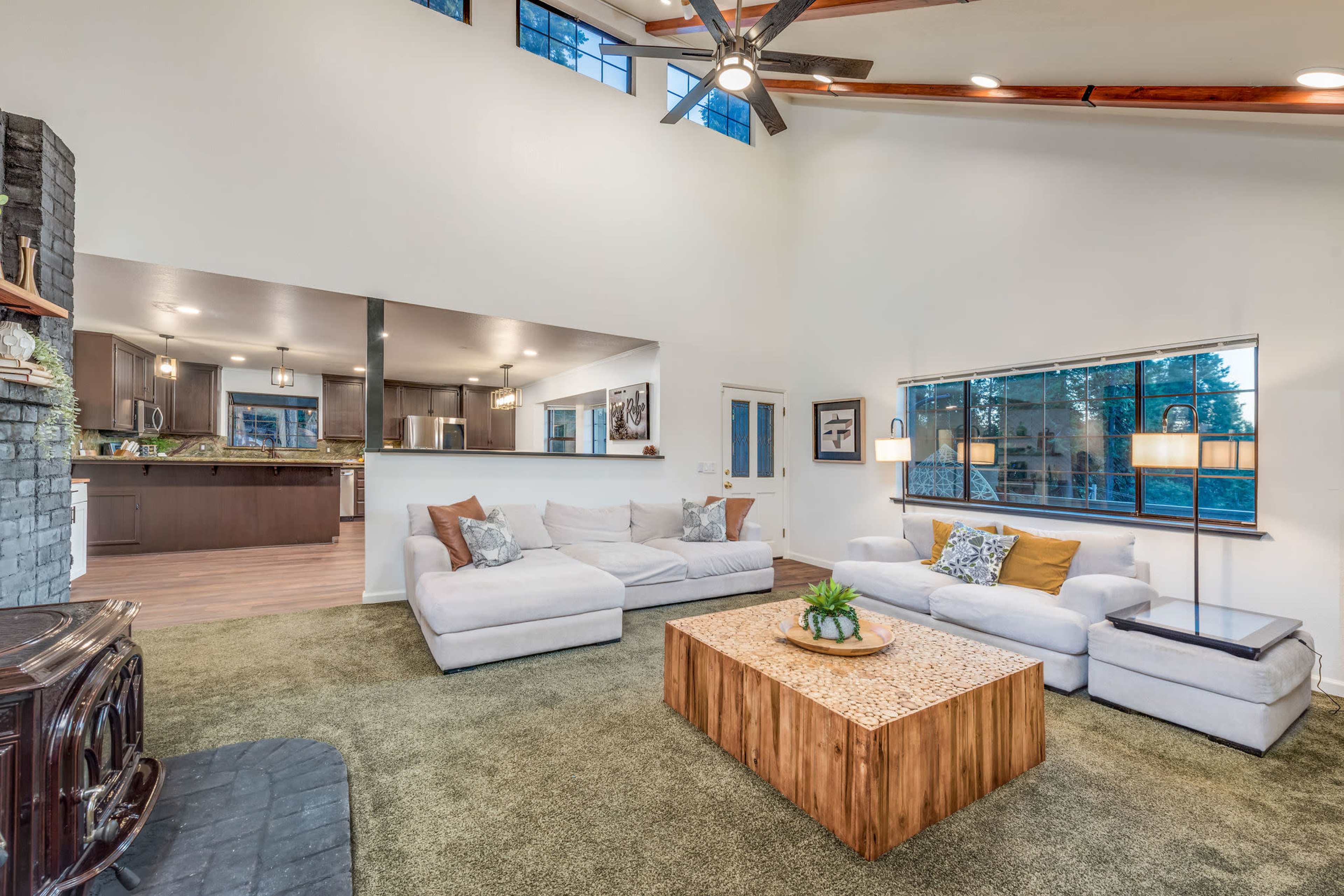 The spacious living room features a light-colored sectional sofa, a wooden coffee table, and large windows that overlook a kitchen area.