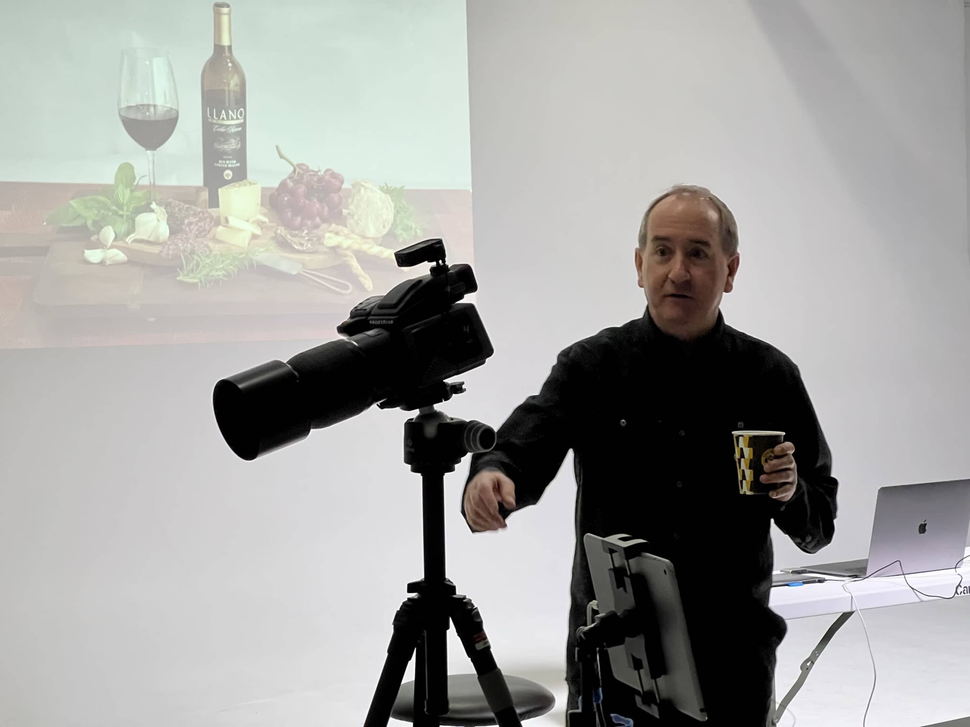 A man stands beside a camera on a tripod, holding a cup, while a still life of food and wine is projected on the wall behind him.