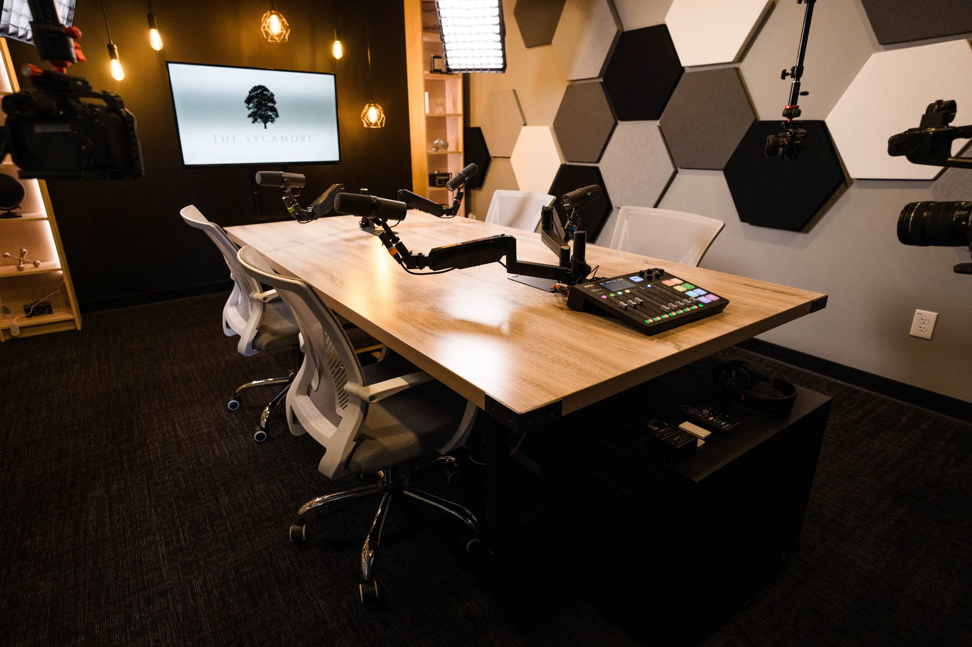 A modern conference room features a large wooden table with cameras, monitors, and a control panel for recording or broadcasting.