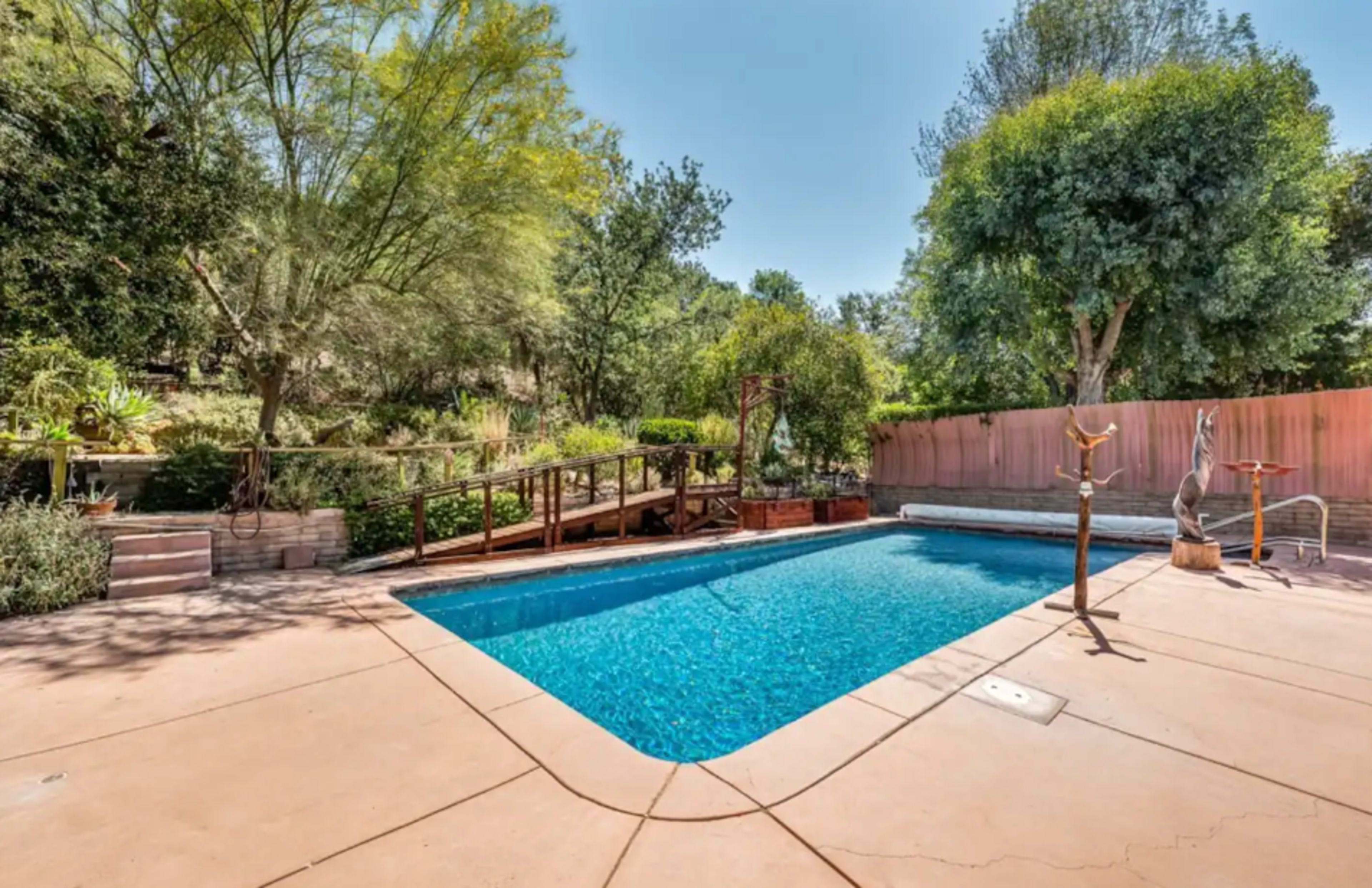 A rectangular swimming pool is surrounded by landscaped greenery and a wooden walkway leads to a garden area in the background.
