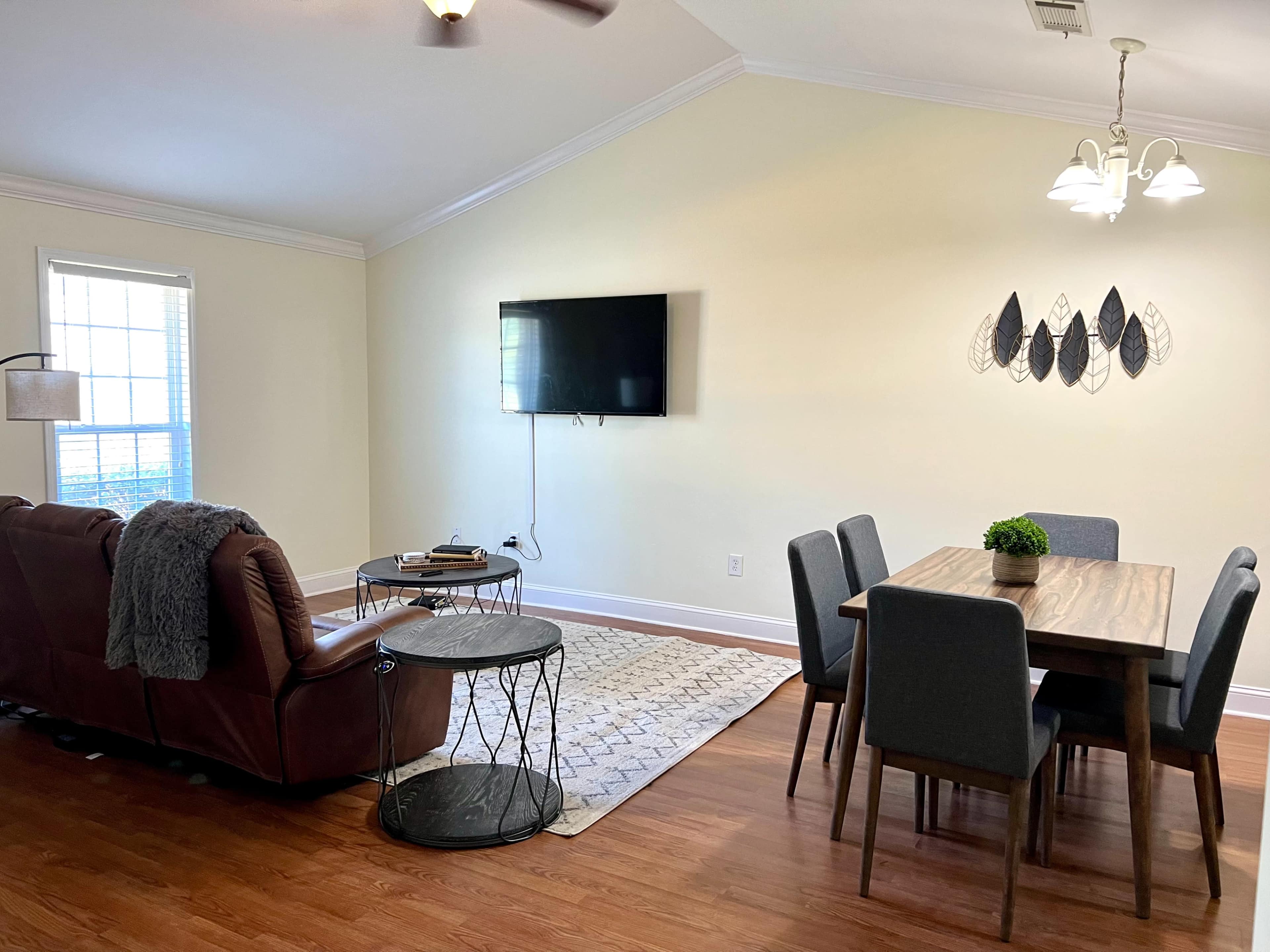 A living space featuring a brown leather sofa, a dining table with gray chairs, a wall-mounted TV, and decorative wall art.