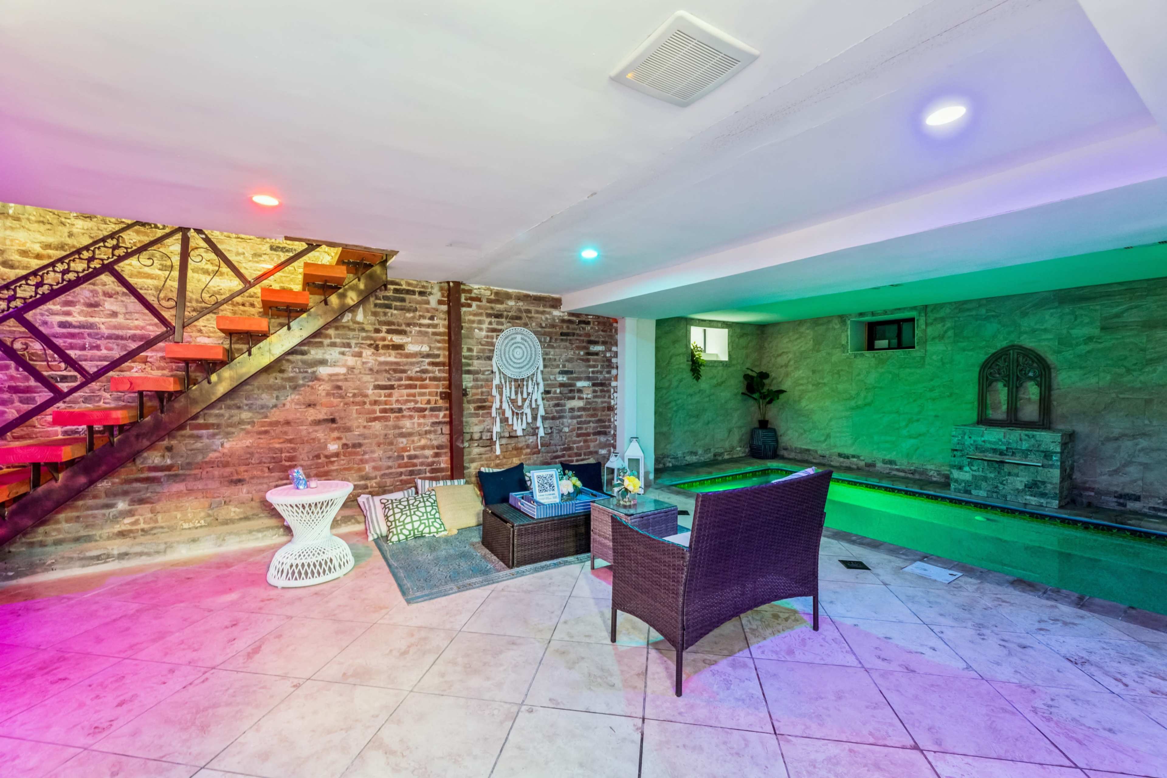 A basement area featuring a staircase with a wooden railing, a seating arrangement, and a small indoor pool with colored lighting.