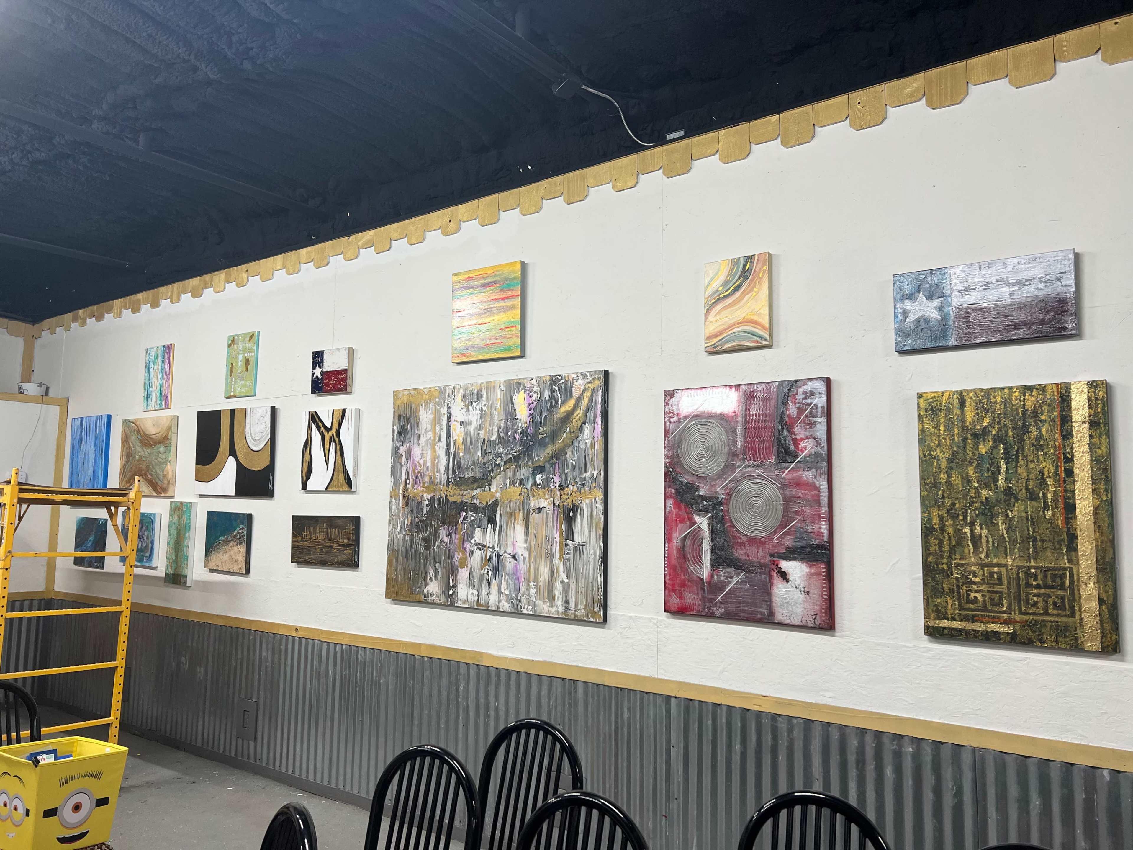 A collection of various paintings displayed on a white wall in a room, with a ladder positioned nearby and black chairs in the foreground.