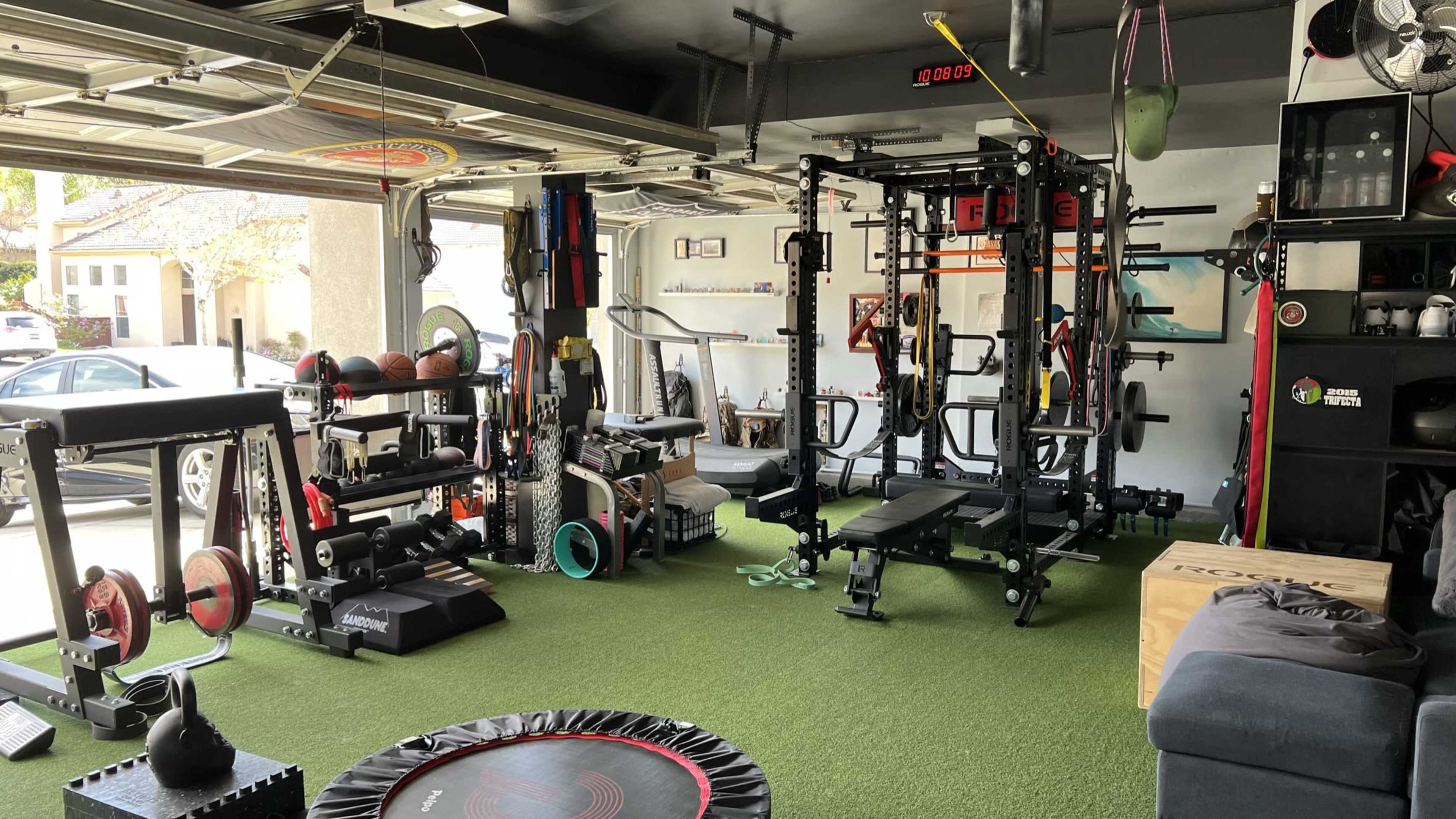 The image shows a home garage gym equipped with various fitness machines, weights, and exercise accessories on a green turf floor.