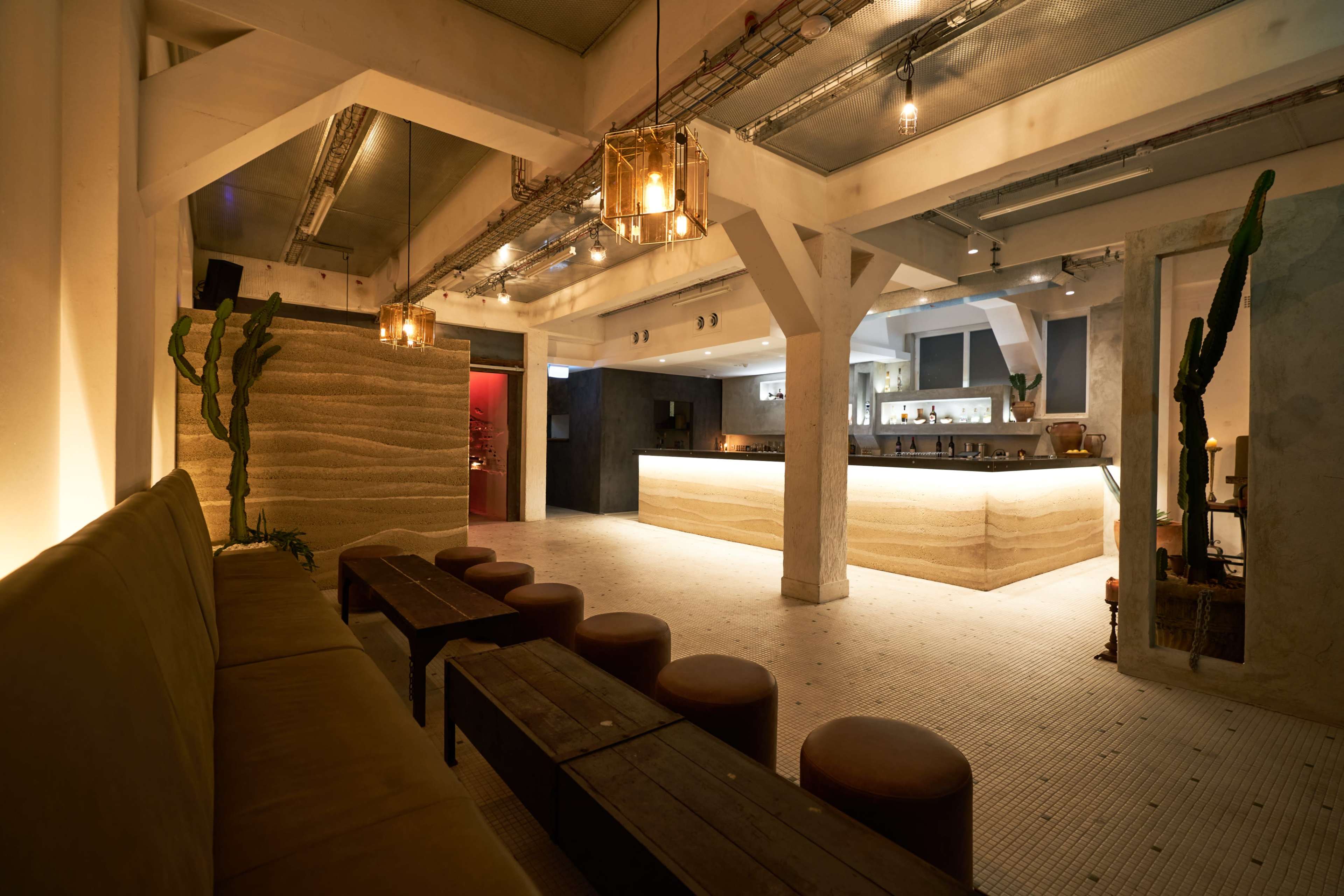 The image shows a modern, minimally furnished bar interior with wooden seating and a well-lit counter.