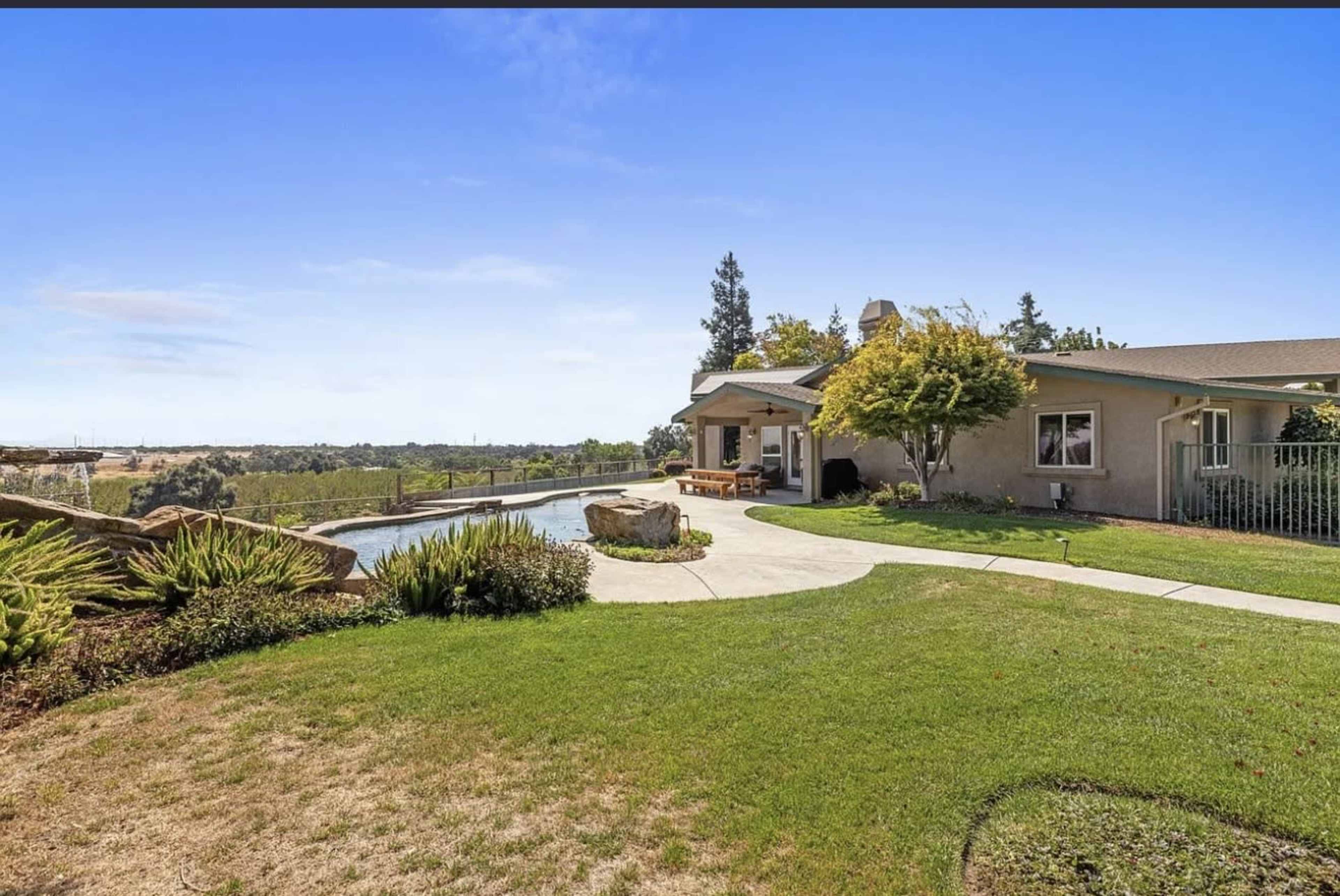 The image shows a single-story house with a landscaped yard, a pool, and a backdrop of green hills under a clear blue sky.