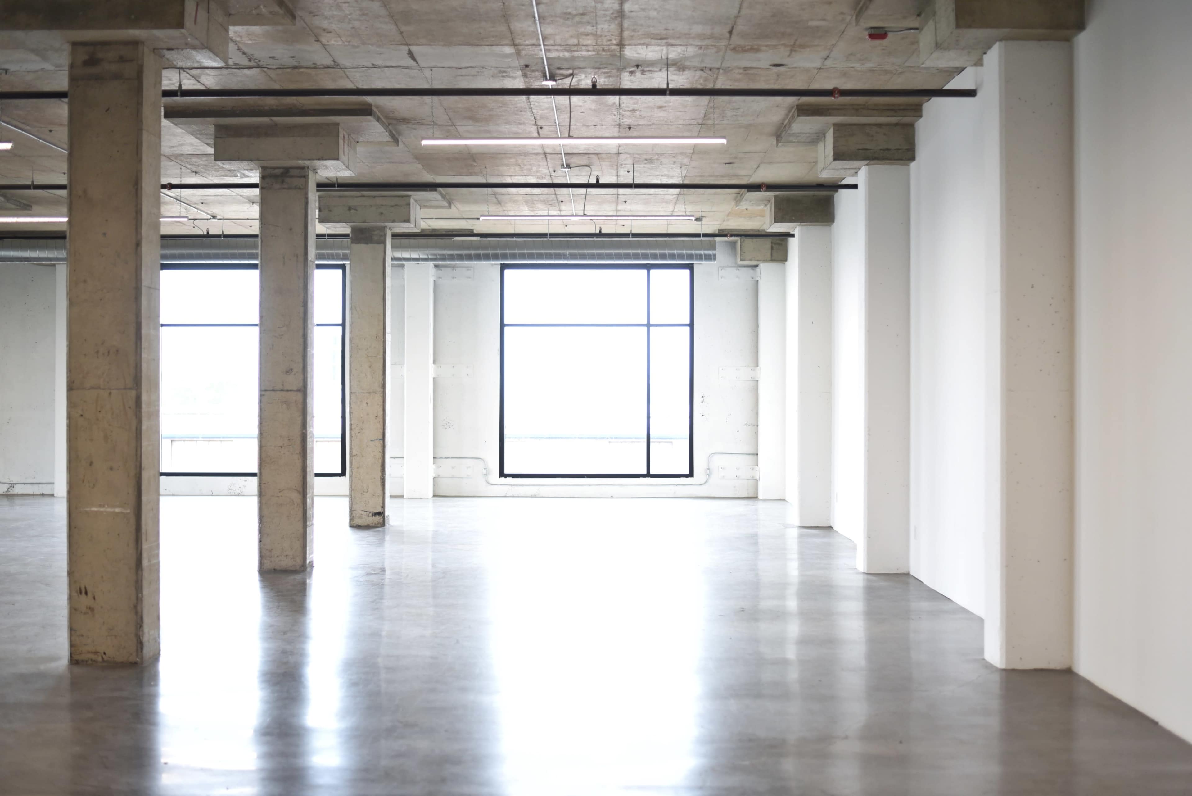 The image shows a spacious, empty industrial-style room with concrete pillars and large windows.