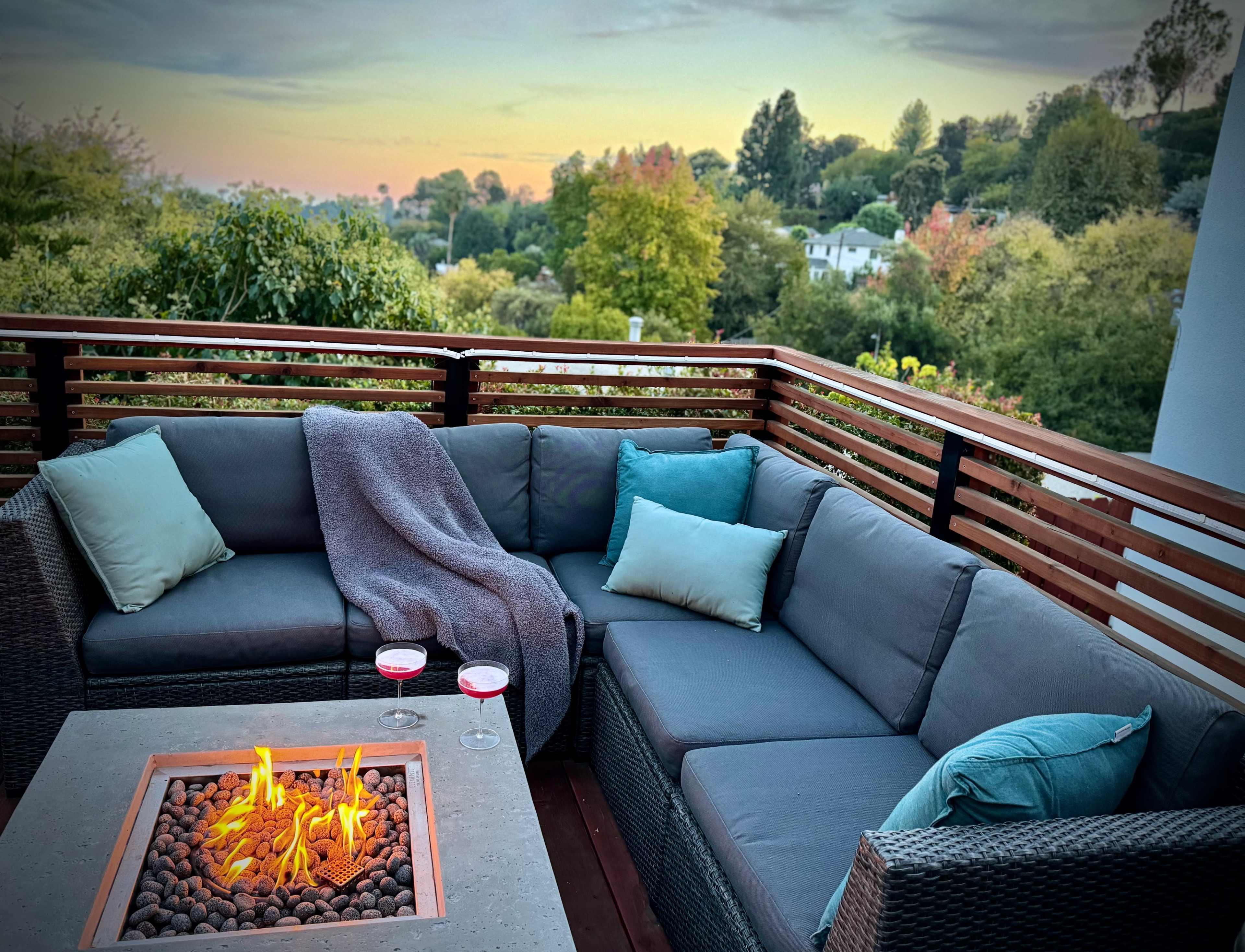 A cozy outdoor seating area features a sectional sofa with cushions, a fire pit with flames, and two cocktails on a nearby table, set against a backdrop of wooded hills at sunset.