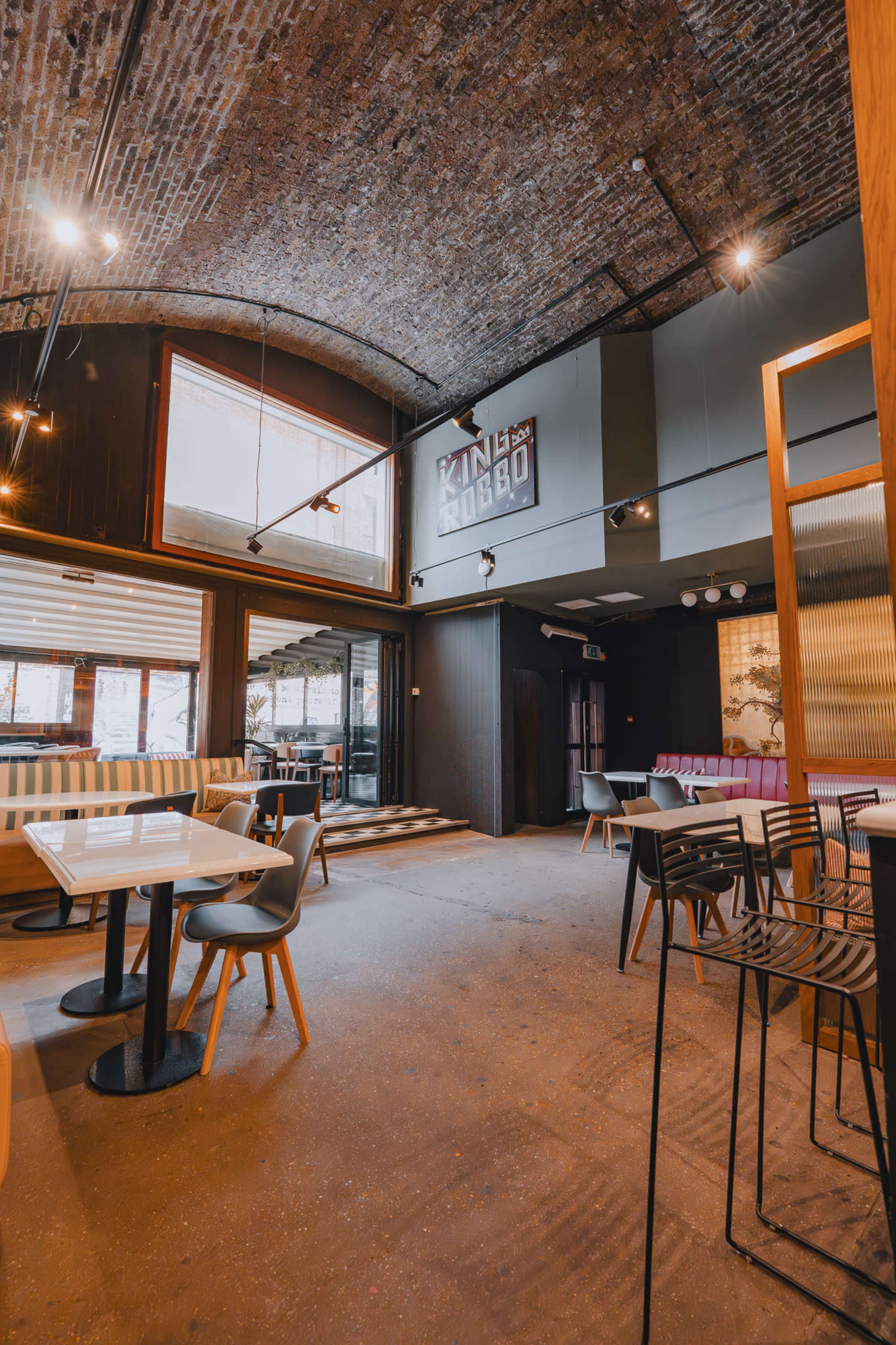 The image shows a modern restaurant interior with a high arched brick ceiling, tables and chairs arranged throughout, and large windows allowing natural light to enter.