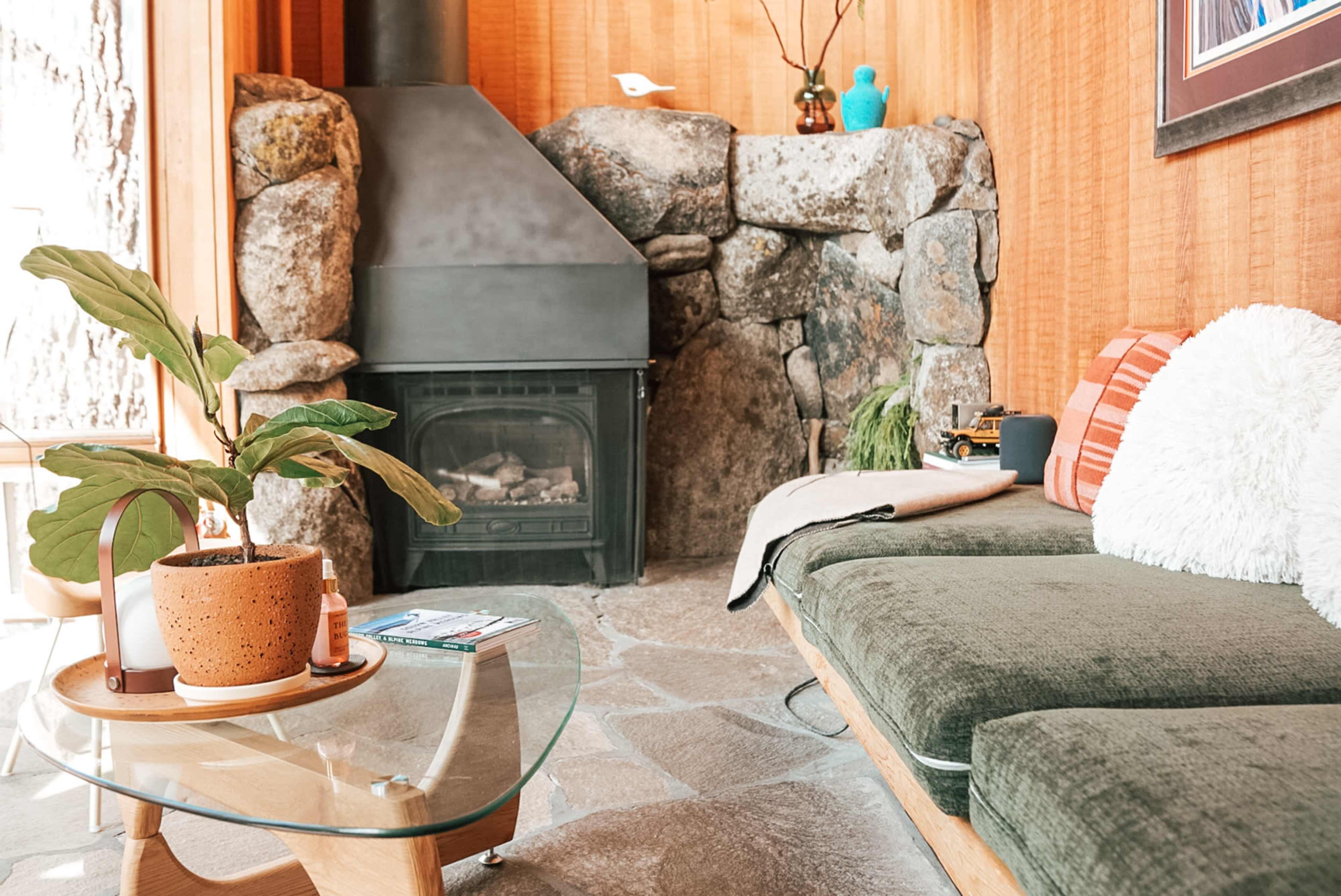 The image shows a cozy living area featuring a stone fireplace, a green sofa, a glass coffee table, and various decorative items against a wooden wall.