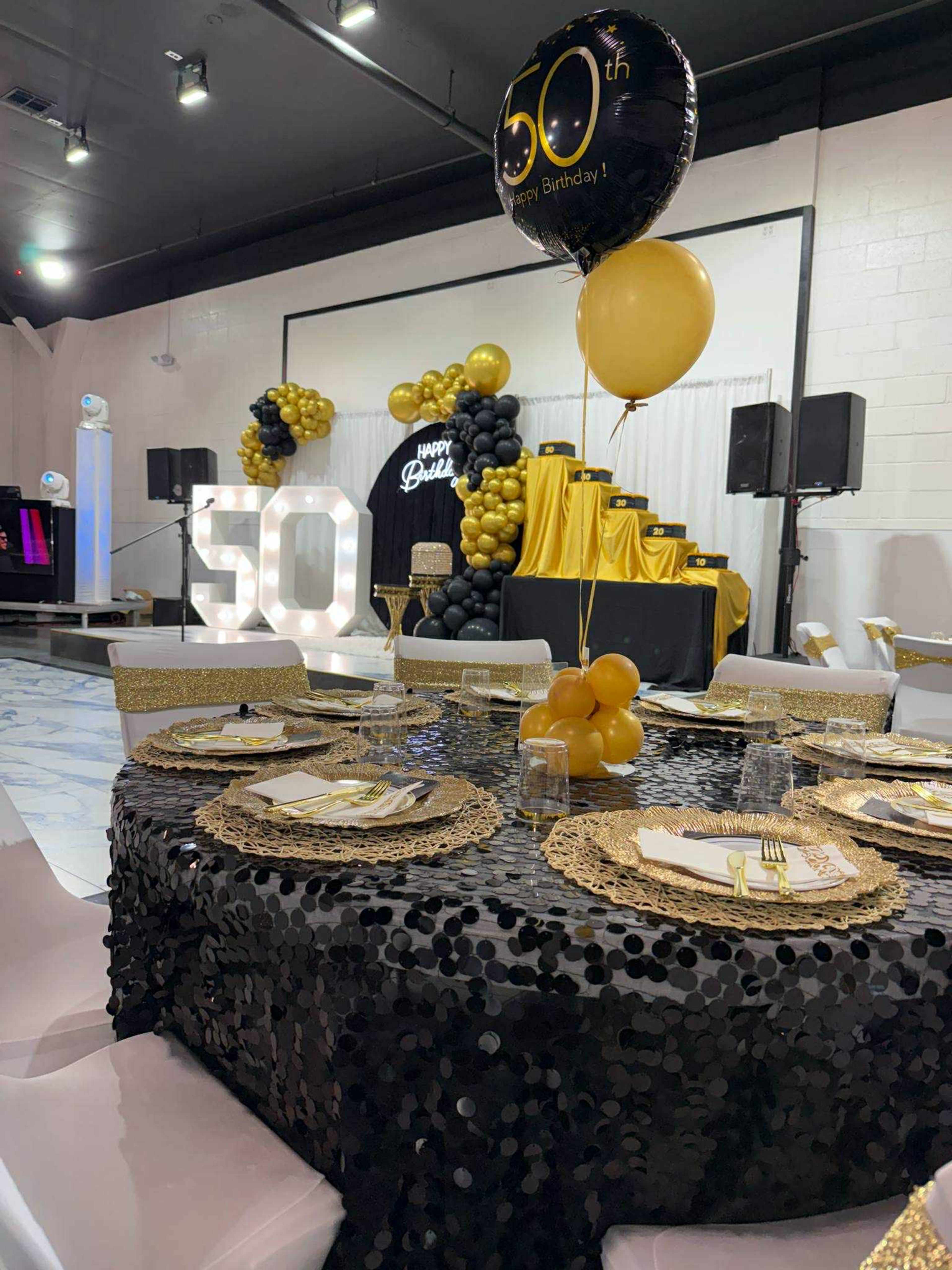 A black and gold-themed birthday party setup features a decorated table with a sparkling tablecloth, gold plates, and a large "50" sign in the background.