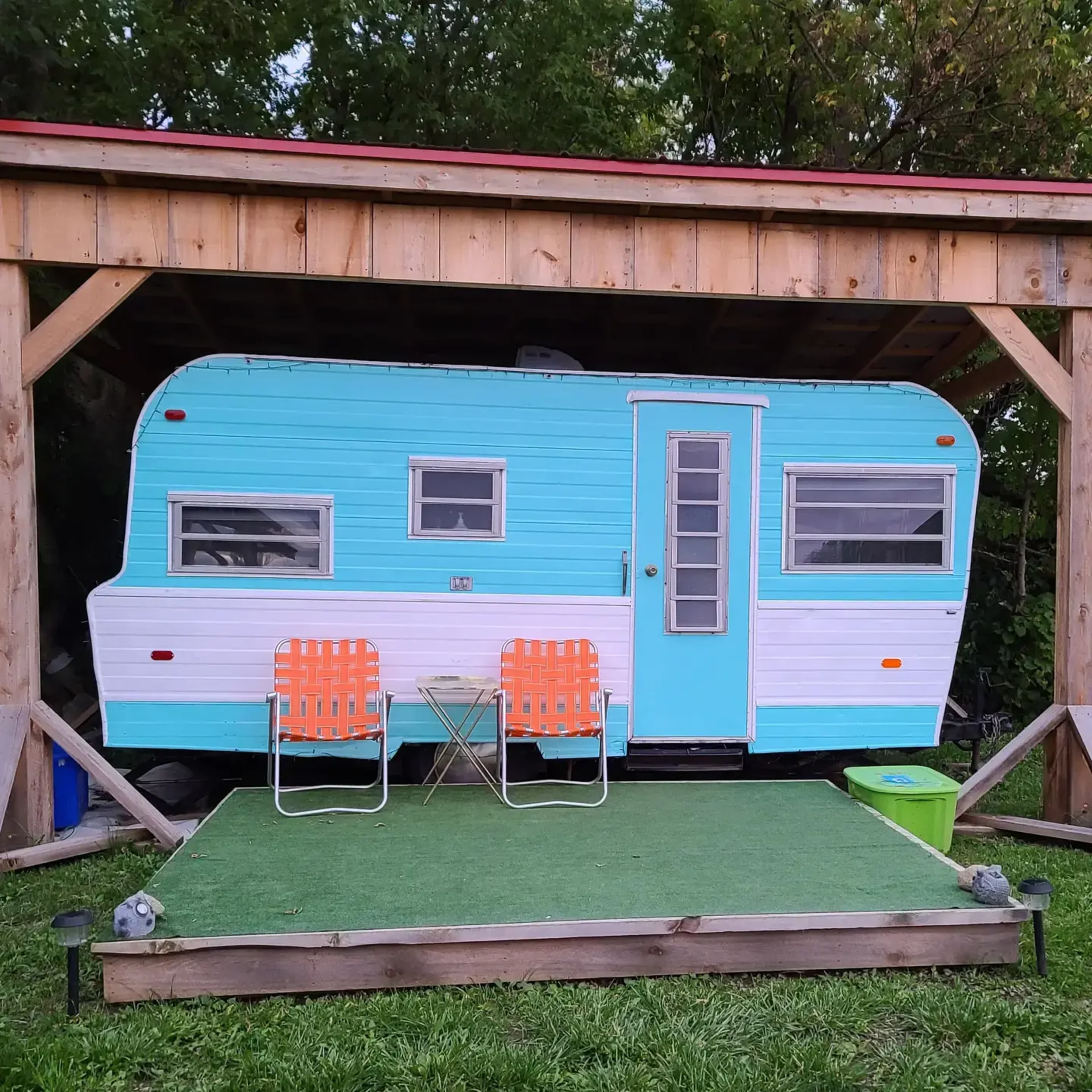 A vintage turquoise and white camper is parked under a wooden shelter, accompanied by two orange chairs and a small table on a green mat.