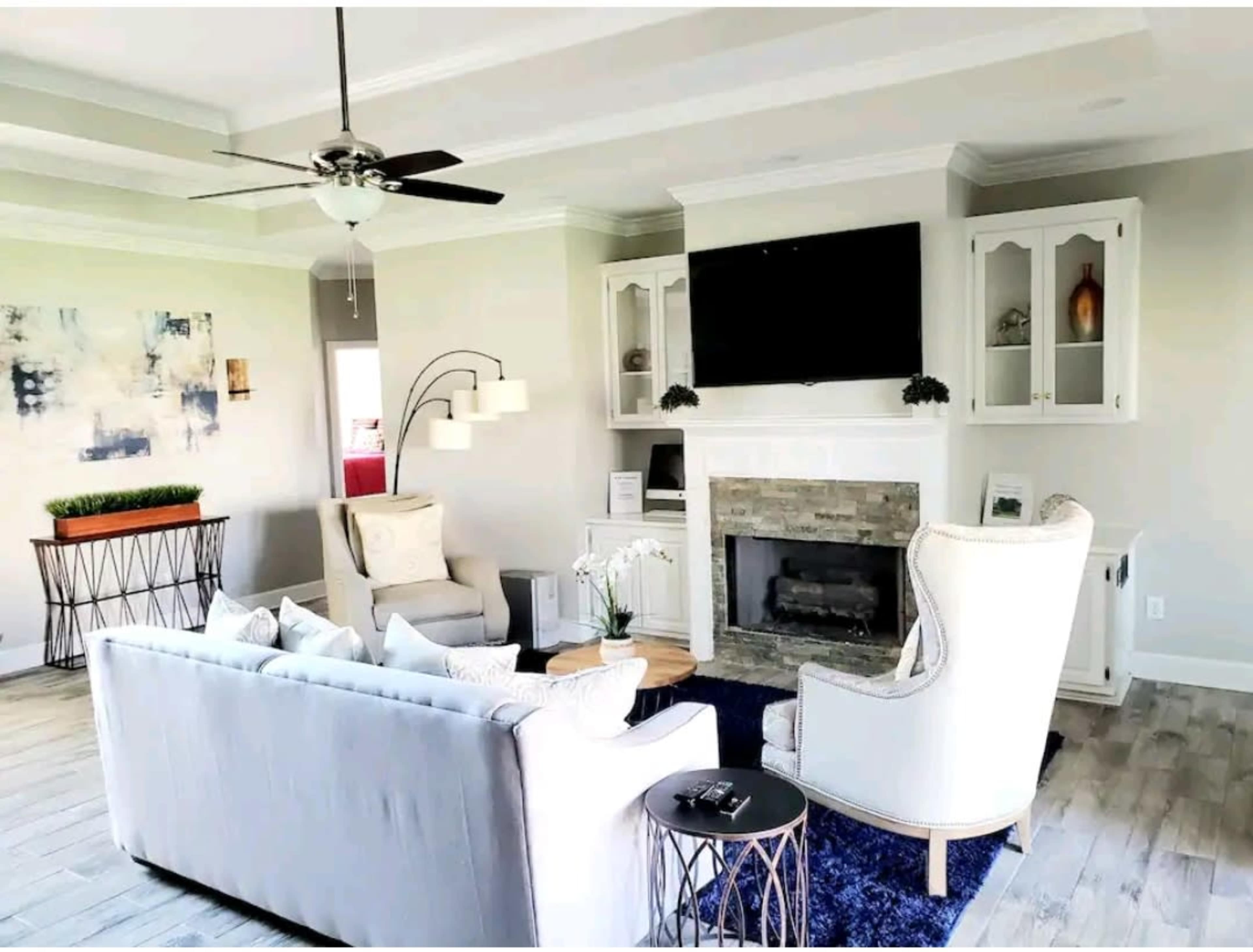 A modern living room features a light-colored sofa, two armchairs, a stone fireplace with a mounted television, and a decorative painting on the wall.