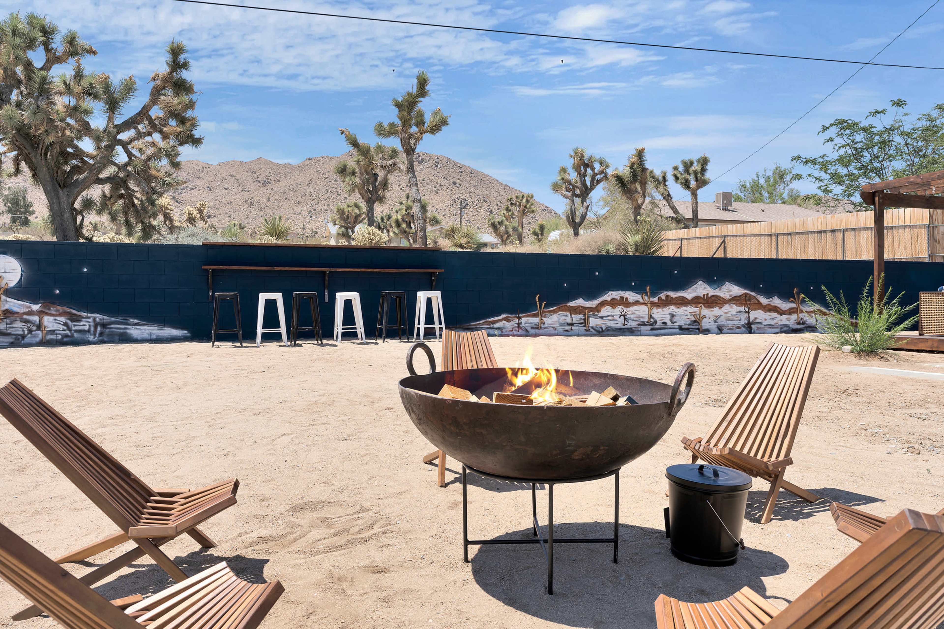 A circular fire pit surrounded by wooden chairs sits in a sandy area with a mural and desert landscape in the background.