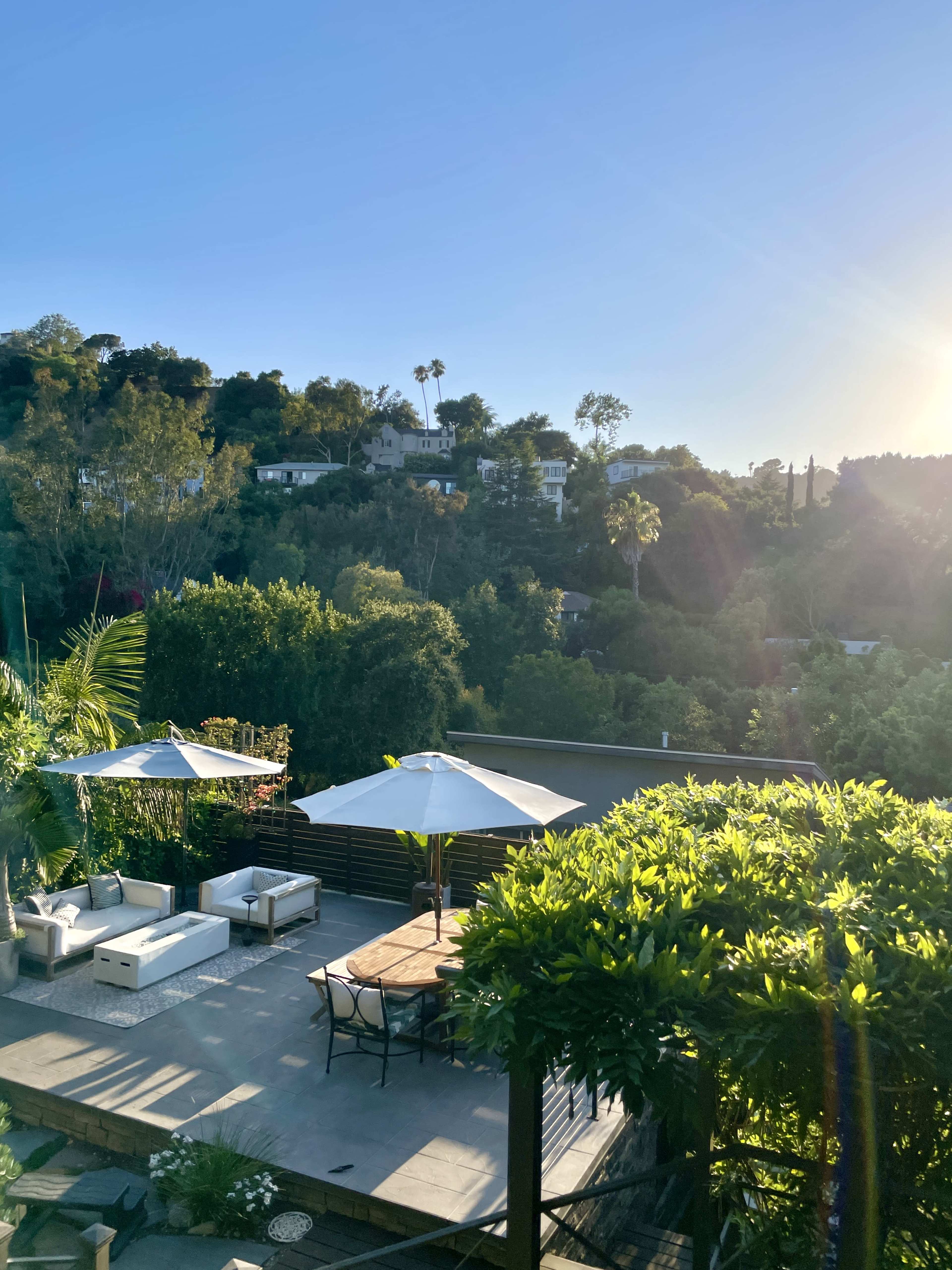 A patio with white outdoor furniture and umbrellas overlooks a hillside with trees and houses under a clear blue sky.