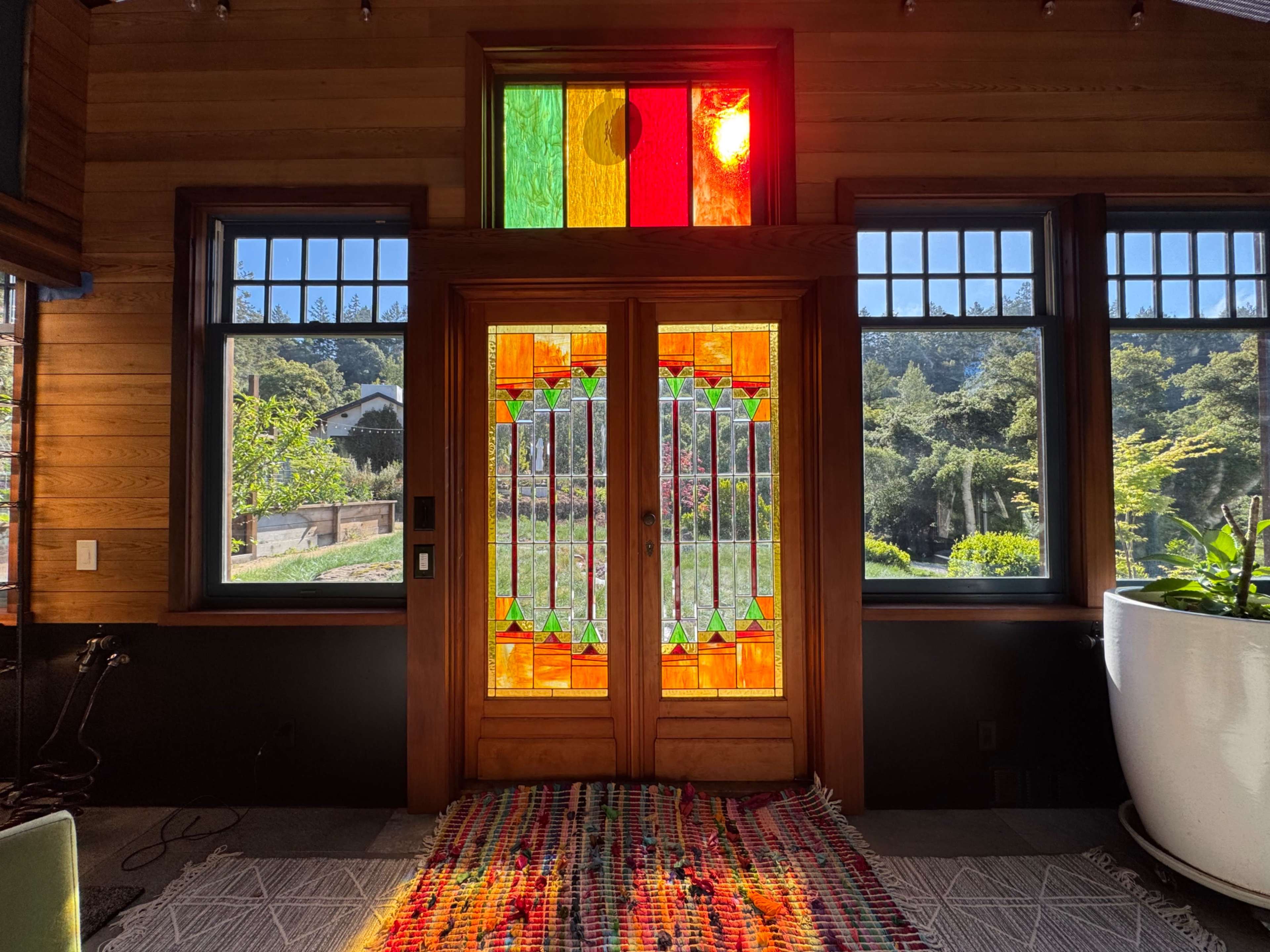 A large stained glass door with colorful panels is illuminated by sunlight, leading to a view of a landscaped garden outside.