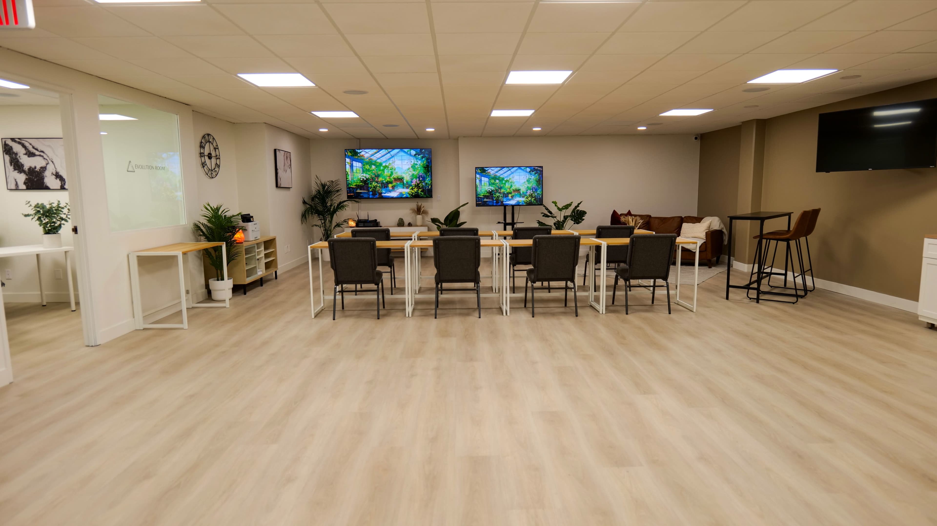 A modern conference room with a long table, several chairs, and two televisions displaying greenery, set in a well-lit space with light wood flooring.