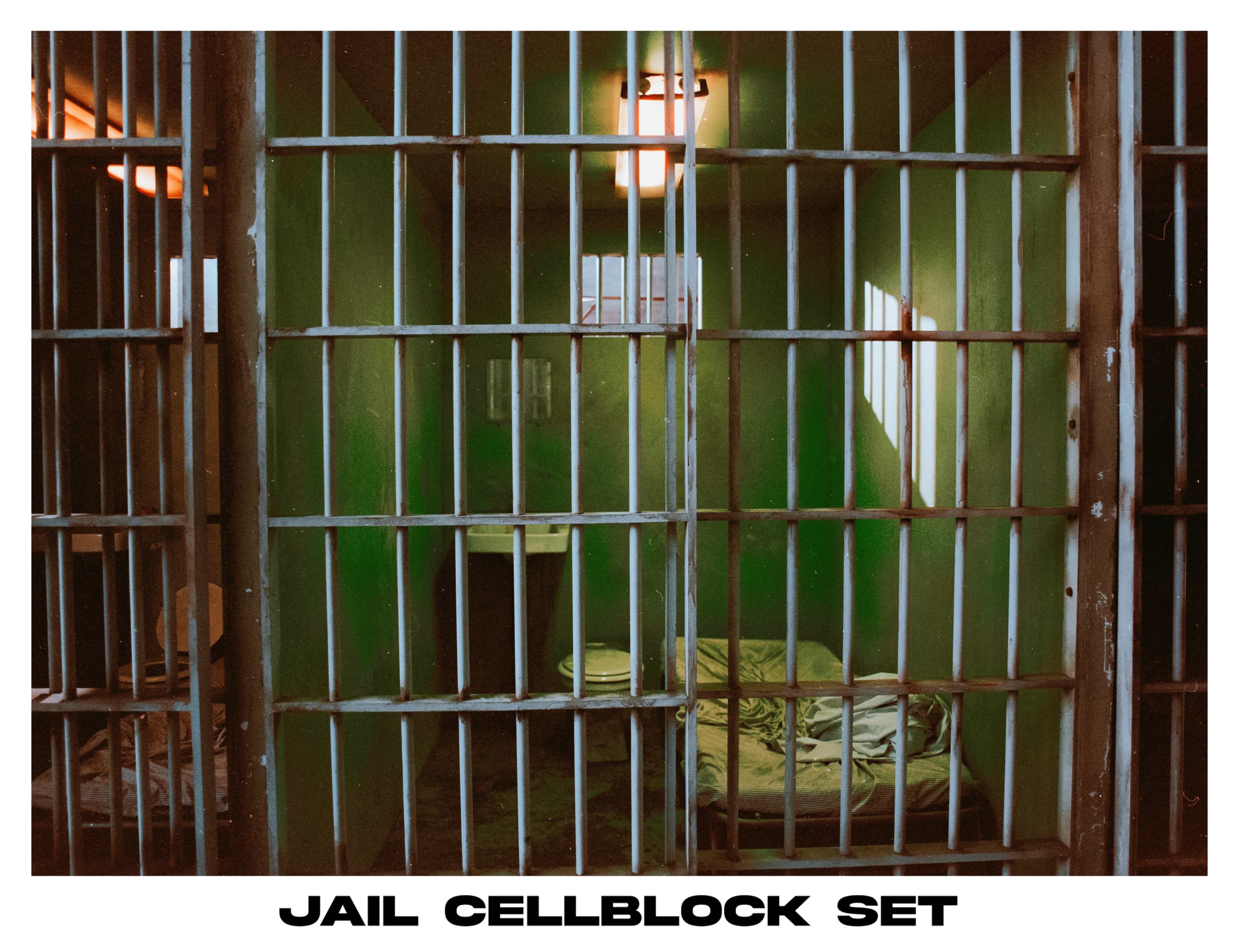 A jail cell block set with barred doors, a bed, and a toilet in a green-painted interior.