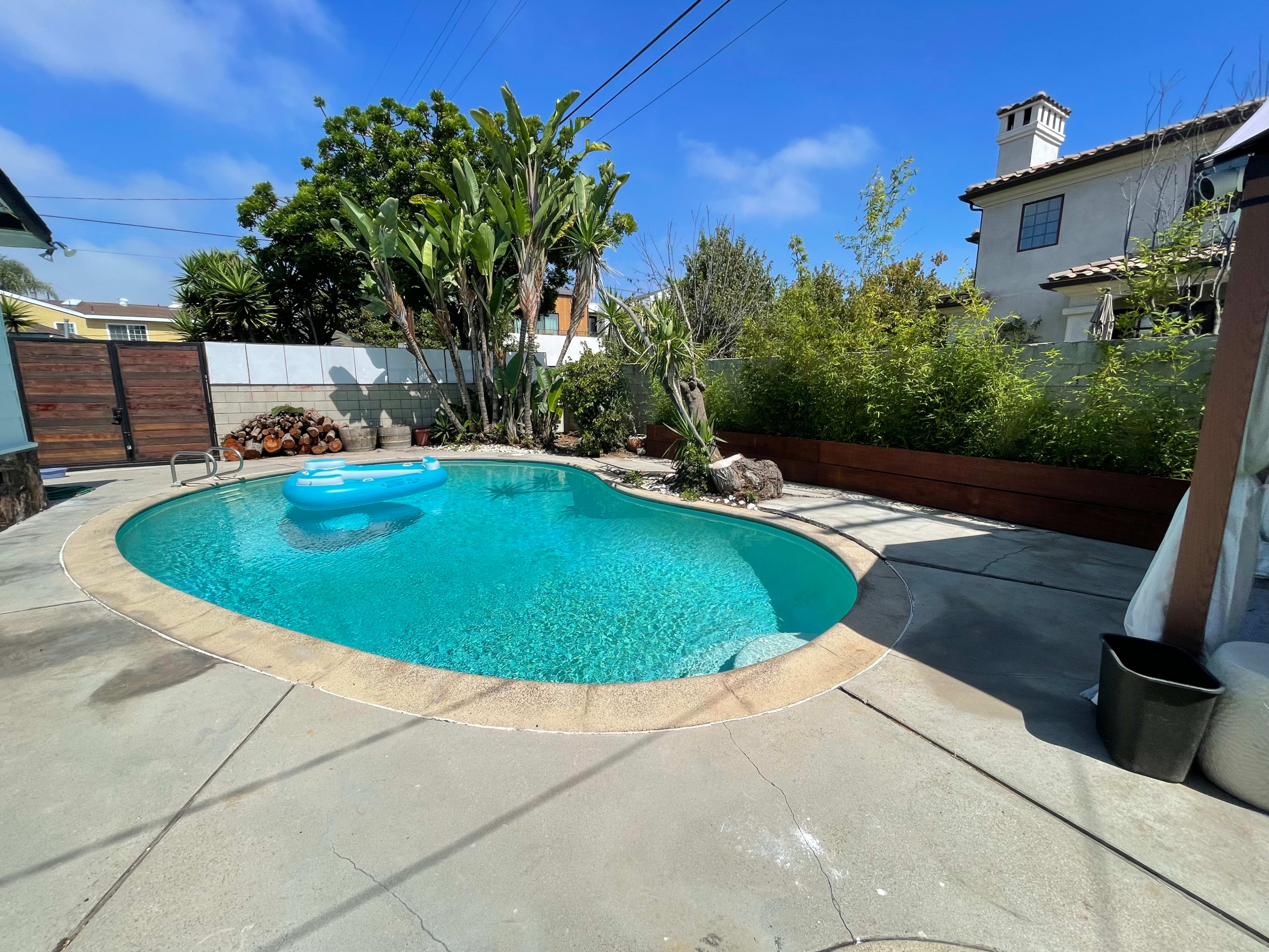 A clear swimming pool is surrounded by tropical plants and stone tiles in a backyard setting.
