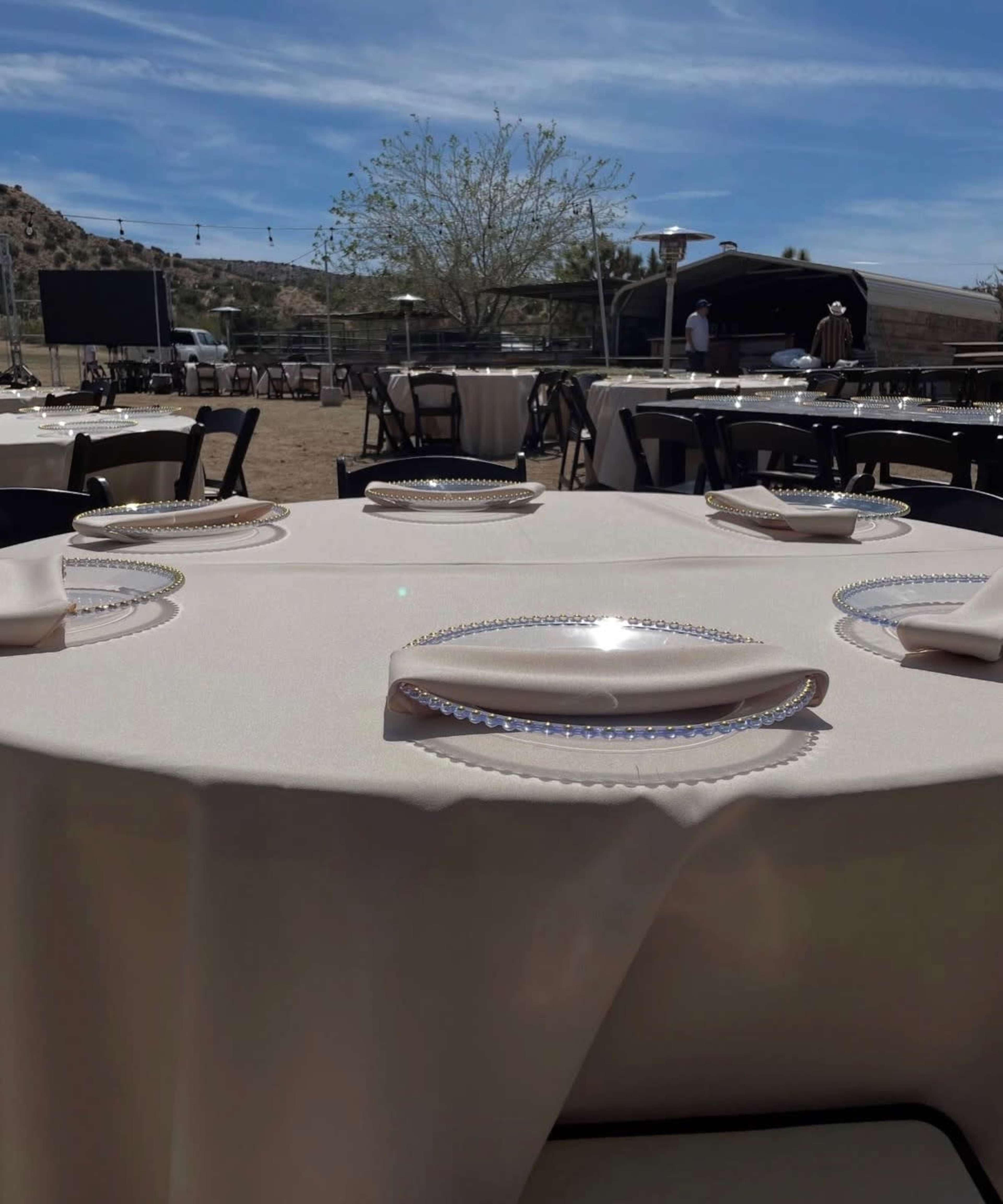 The image shows several tables set outdoors with cream-colored tablecloths and clear plates, under a clear blue sky.