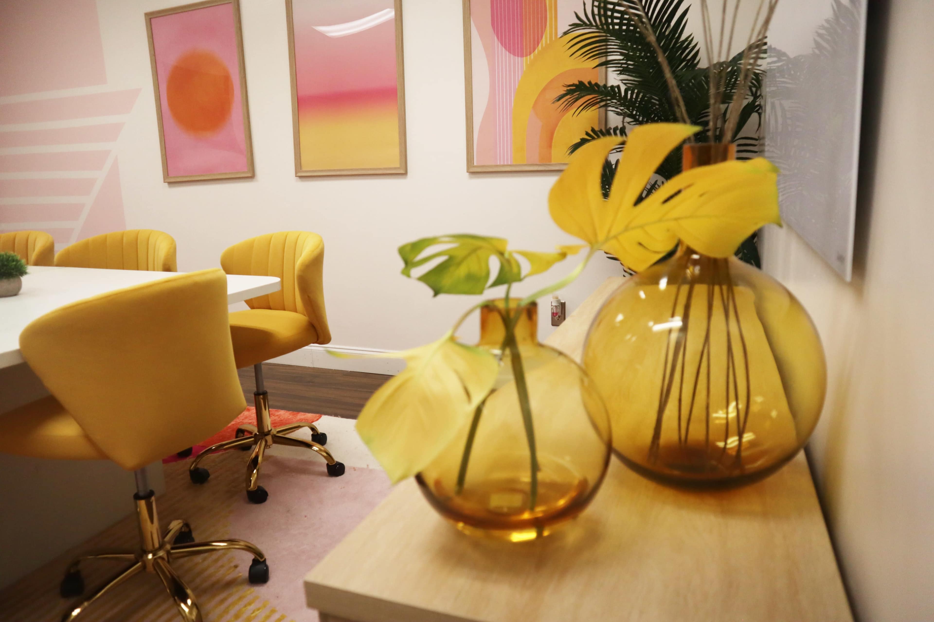 A modern meeting room with yellow chairs, decorative plants in amber vases, and abstract art on the walls.