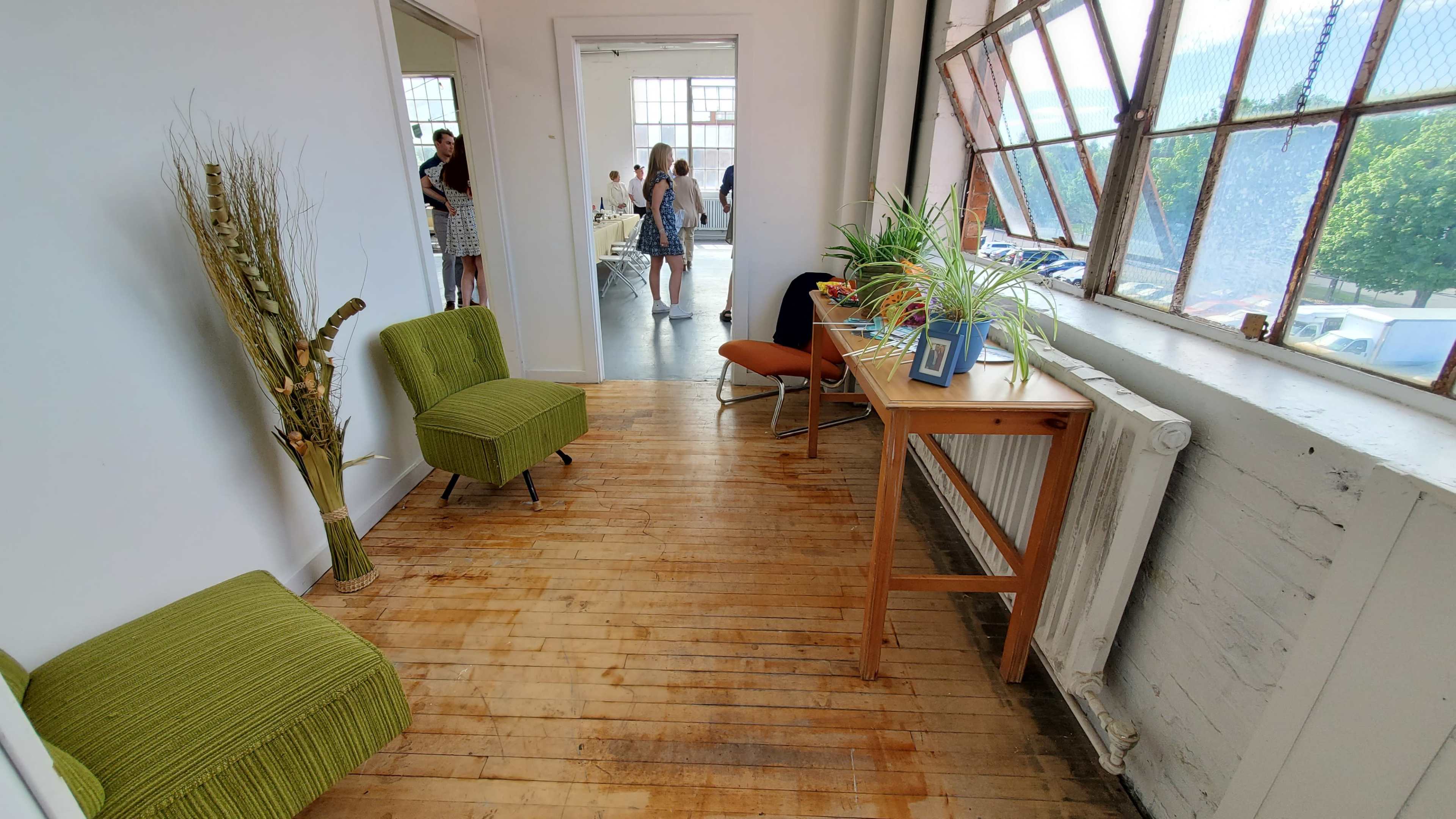 A bright corridor with green chairs, a wooden table, and large windows allowing natural light to fill the space.