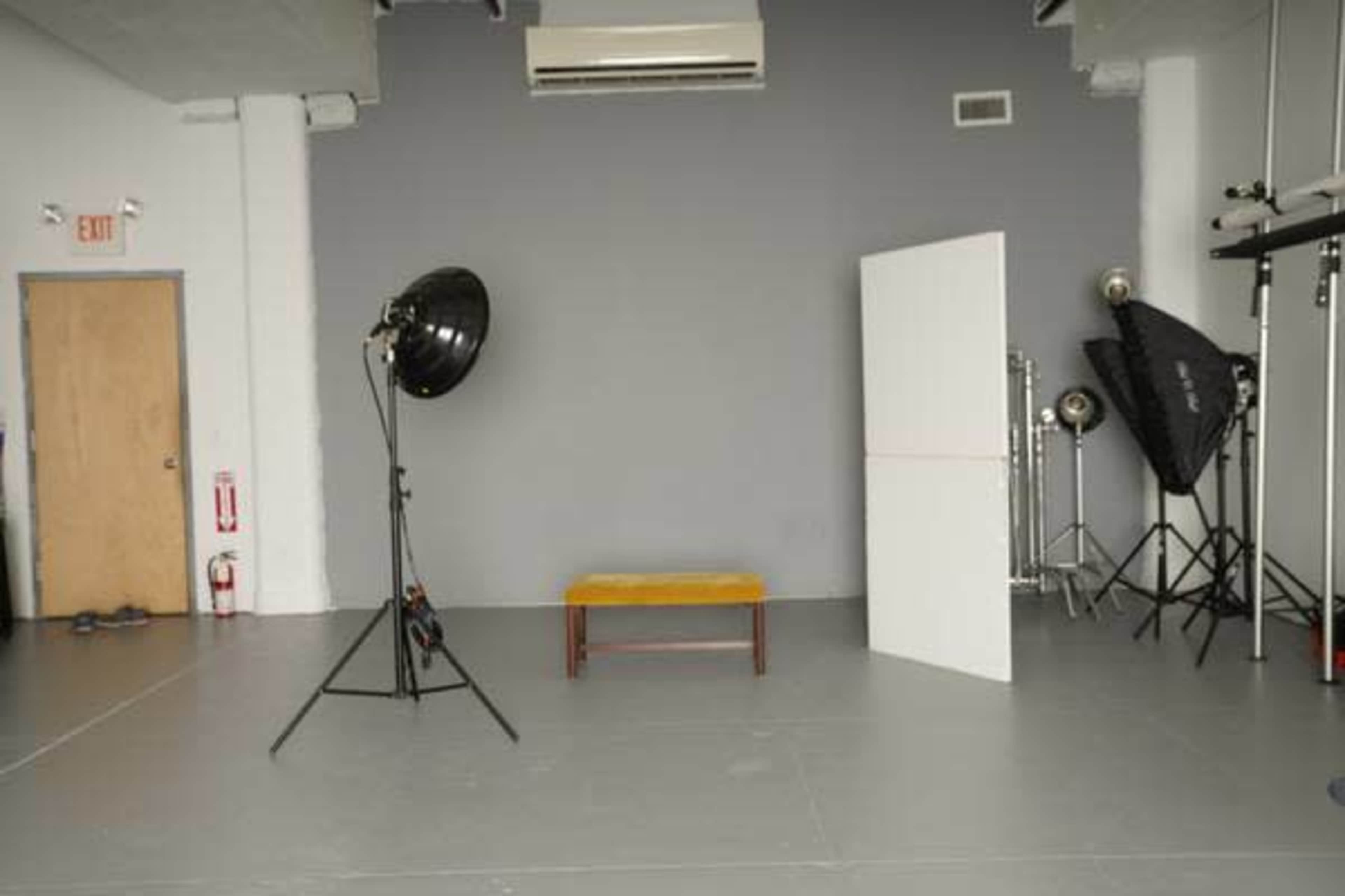 A photography studio with a gray backdrop, a wooden bench in the center, and several lighting equipment arranged around the space.