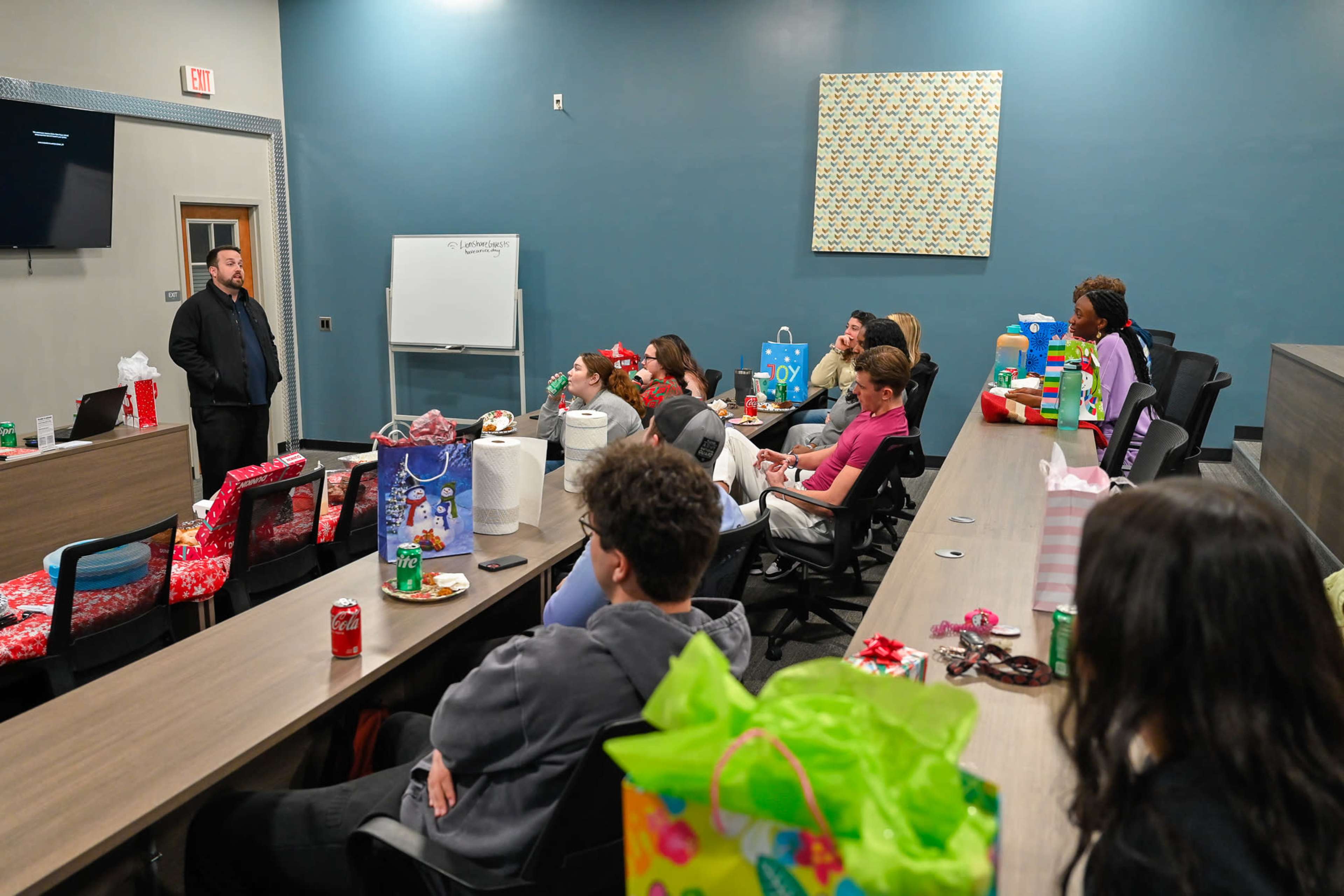 A speaker stands at the front of a conference room filled with people sitting at tables adorned with holiday-themed decorations and gift bags.