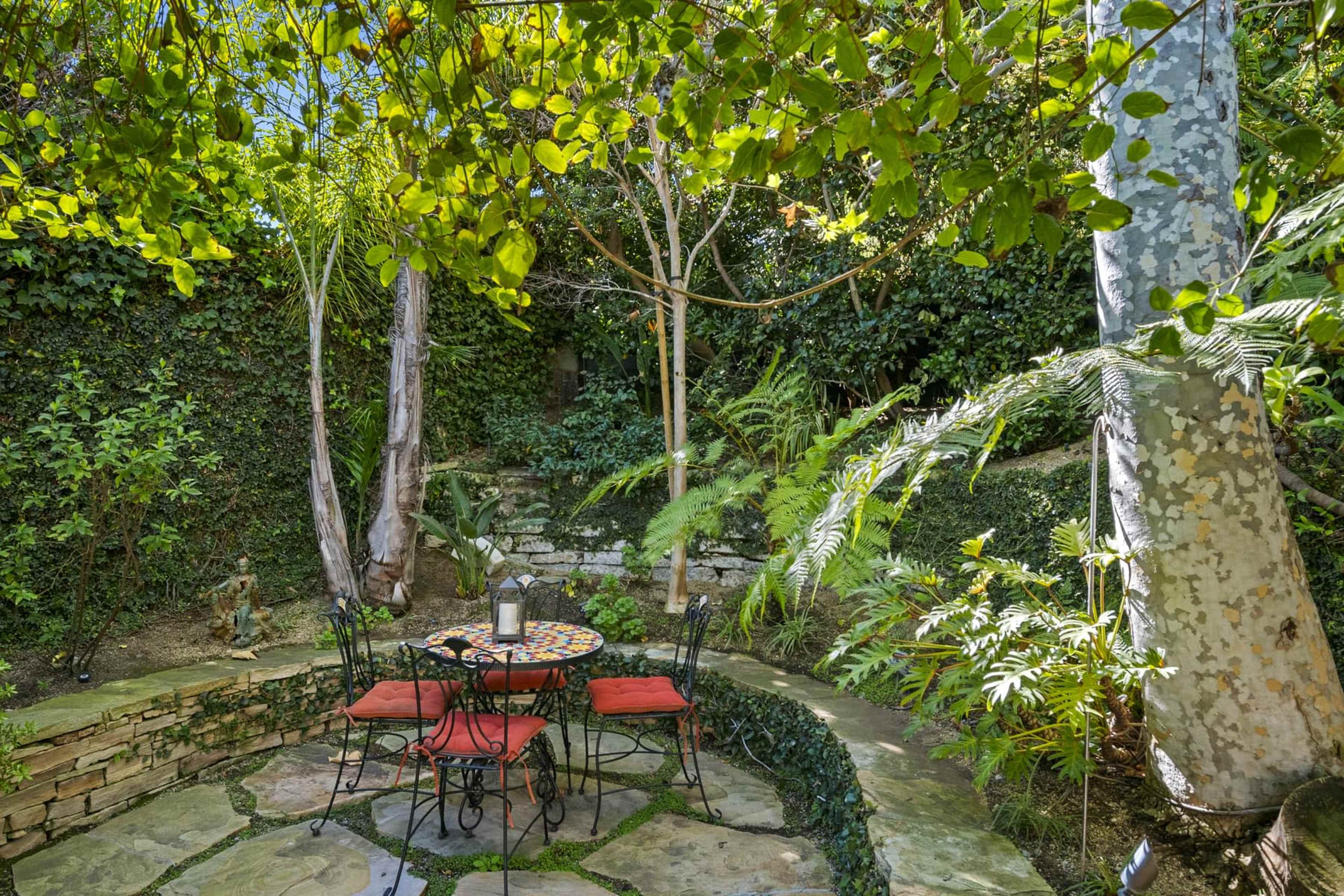 A circular stone patio features a wrought-iron table and chairs surrounded by lush greenery and trees.