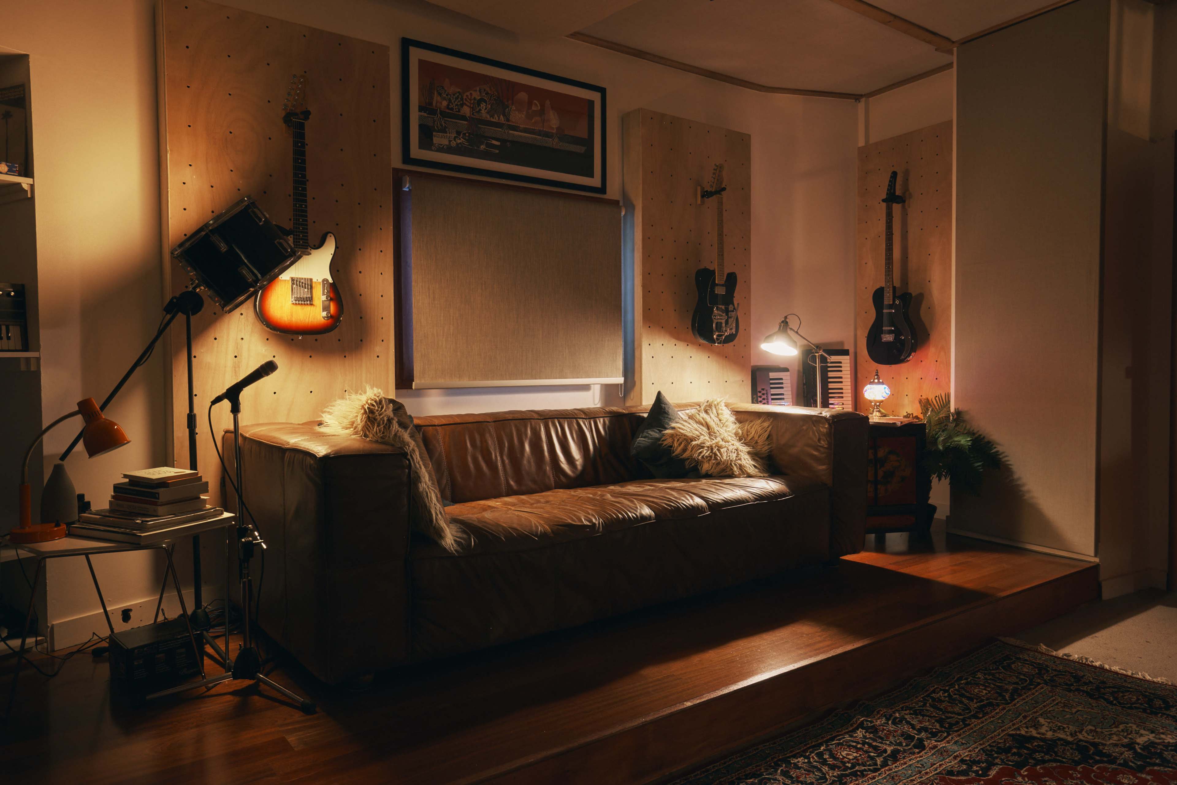 A leather sofa is positioned in a dimly lit room adorned with guitars on wooden panels and a warm desk lamp beside a stack of books.