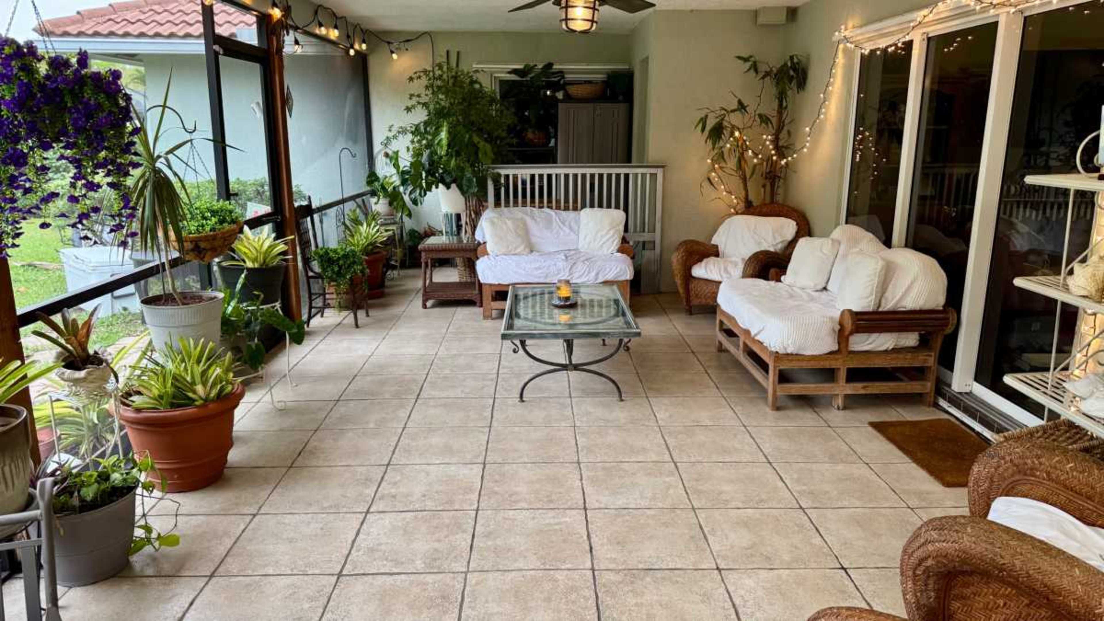 The image shows a screened-in patio decorated with potted plants, wicker furniture, and string lights.
