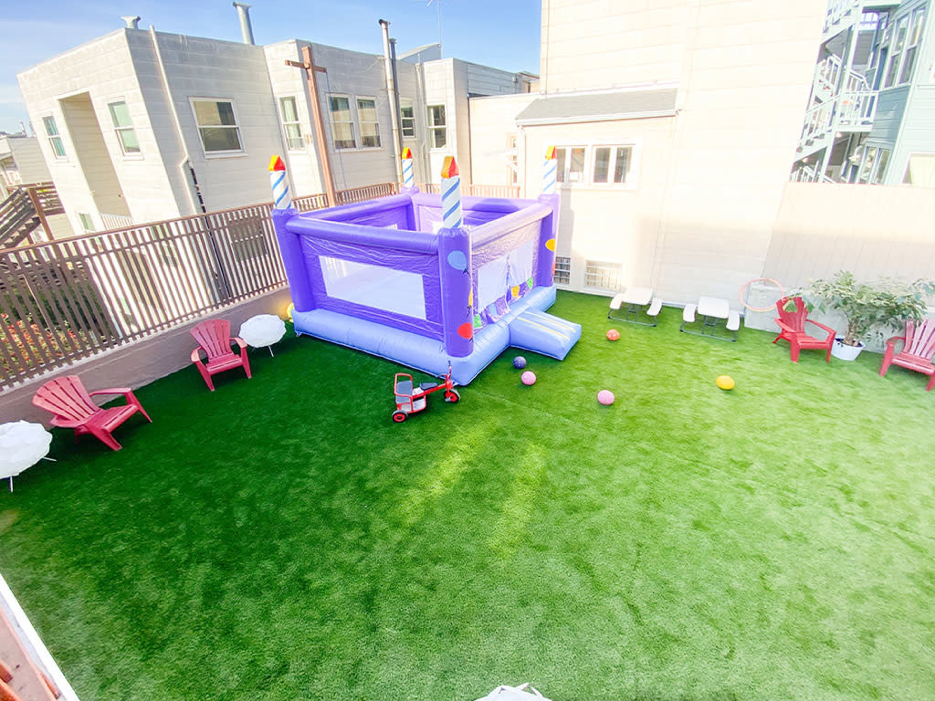 A backyard featuring a purple inflatable bounce house, red Adirondack chairs, colorful balls scattered on the grass, and a small red wagon.